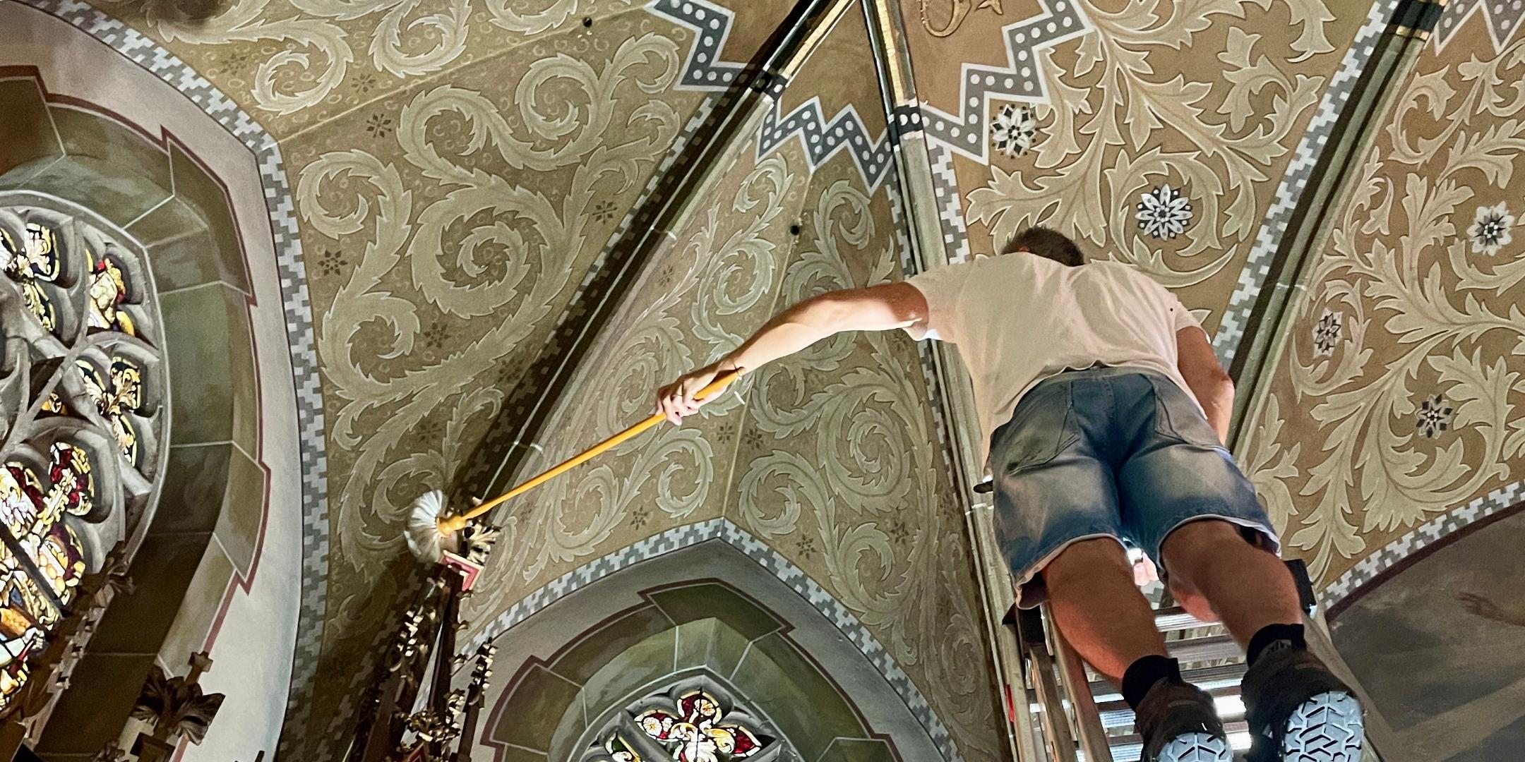 Ein Mann steht in einer Kirche auf einer Leiter vor einem bunten Glasfenster.