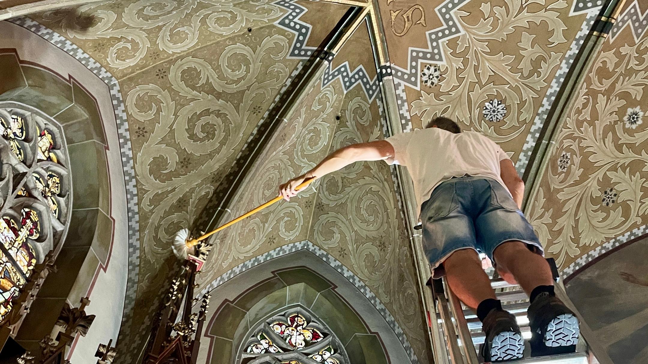 Ein Mann steht in einer Kirche auf einer Leiter vor einem bunten Glasfenster.