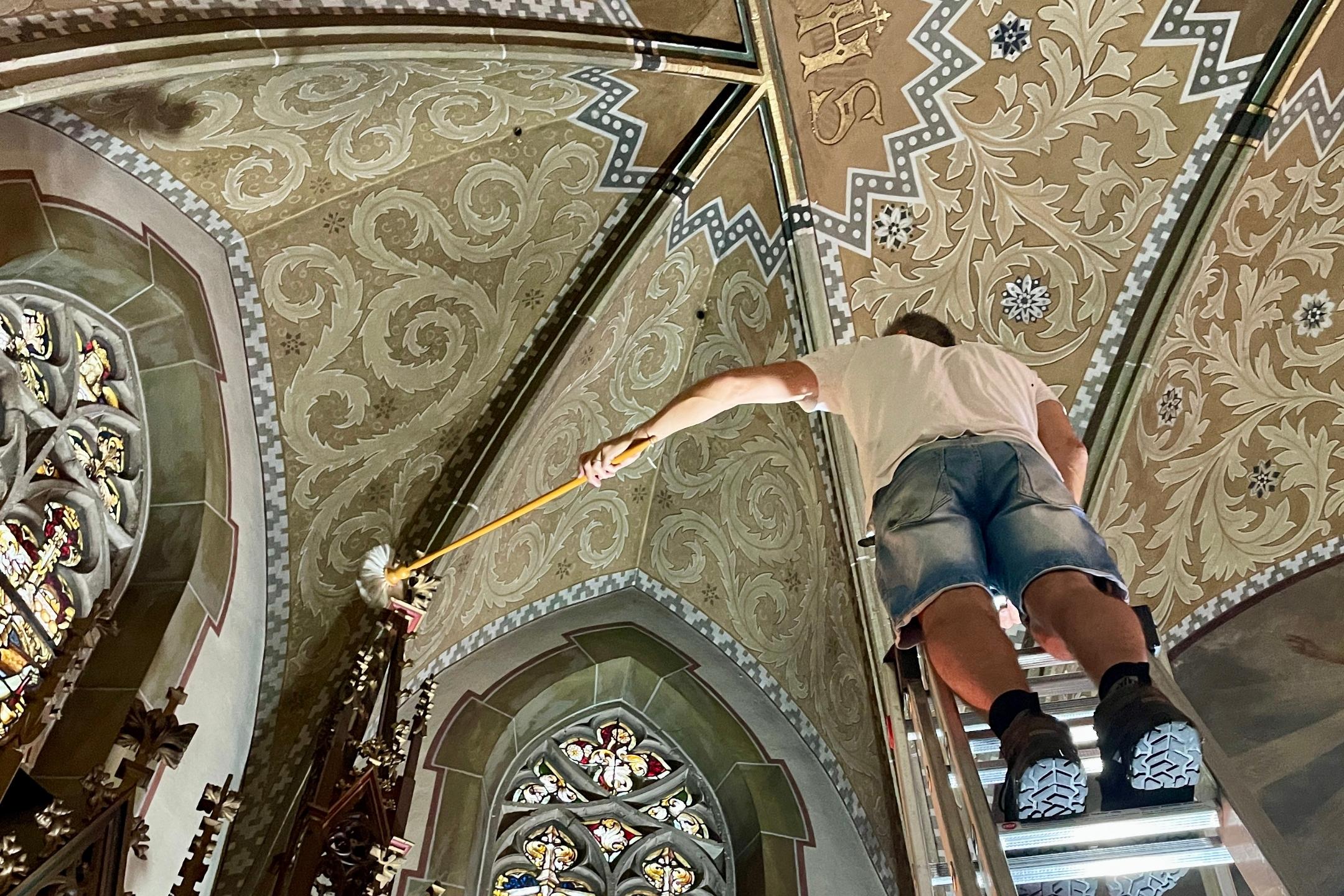 Ein Mann steht in einer Kirche auf einer Leiter vor einem bunten Glasfenster.