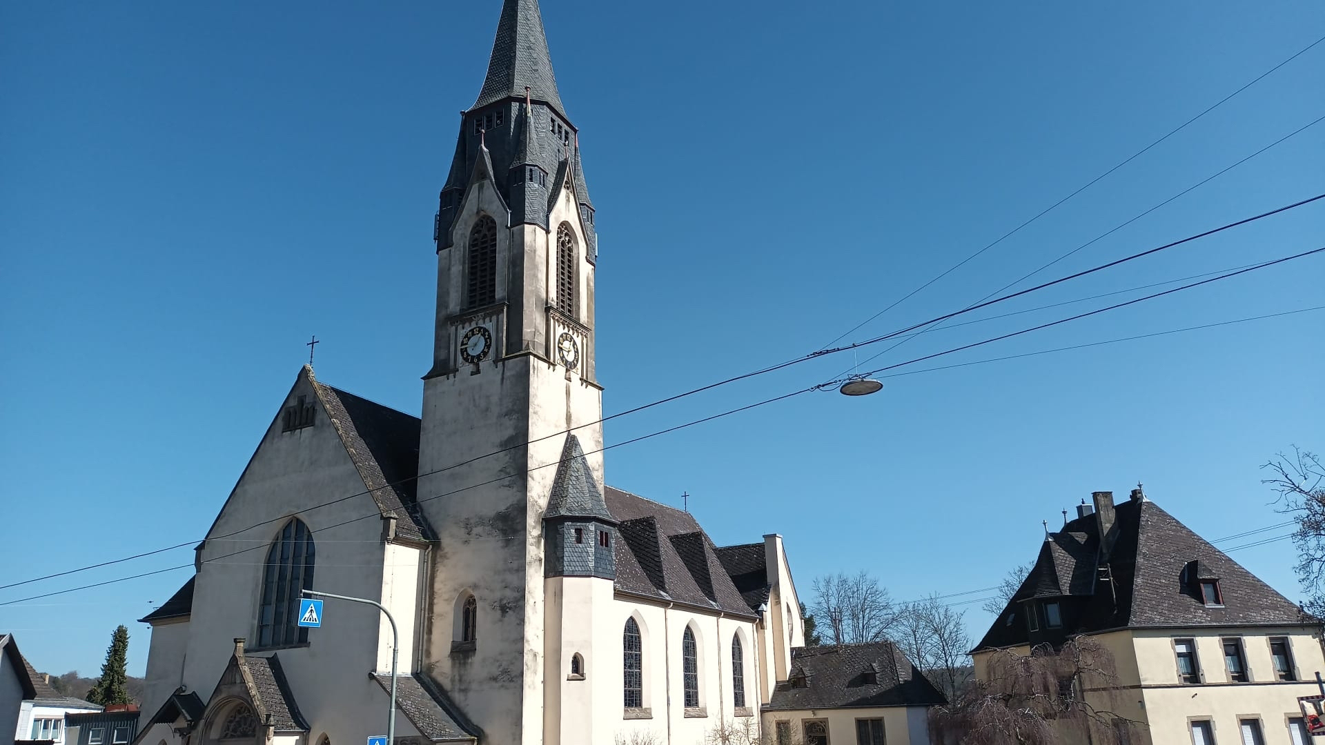 Eine helle, historische Kirche mit hohem, spitzem Glockenturm steht unter klarem blauem Himmel. Das Gebäude zeigt große Kirchenfenster, seitliche Anbauten und ein dunkles Schieferdach. Im Vordergrund verlaufen Strom- und Oberleitungen sowie ein Zebrastreifen. Rechts und links stehen angrenzende Wohnhäuser.