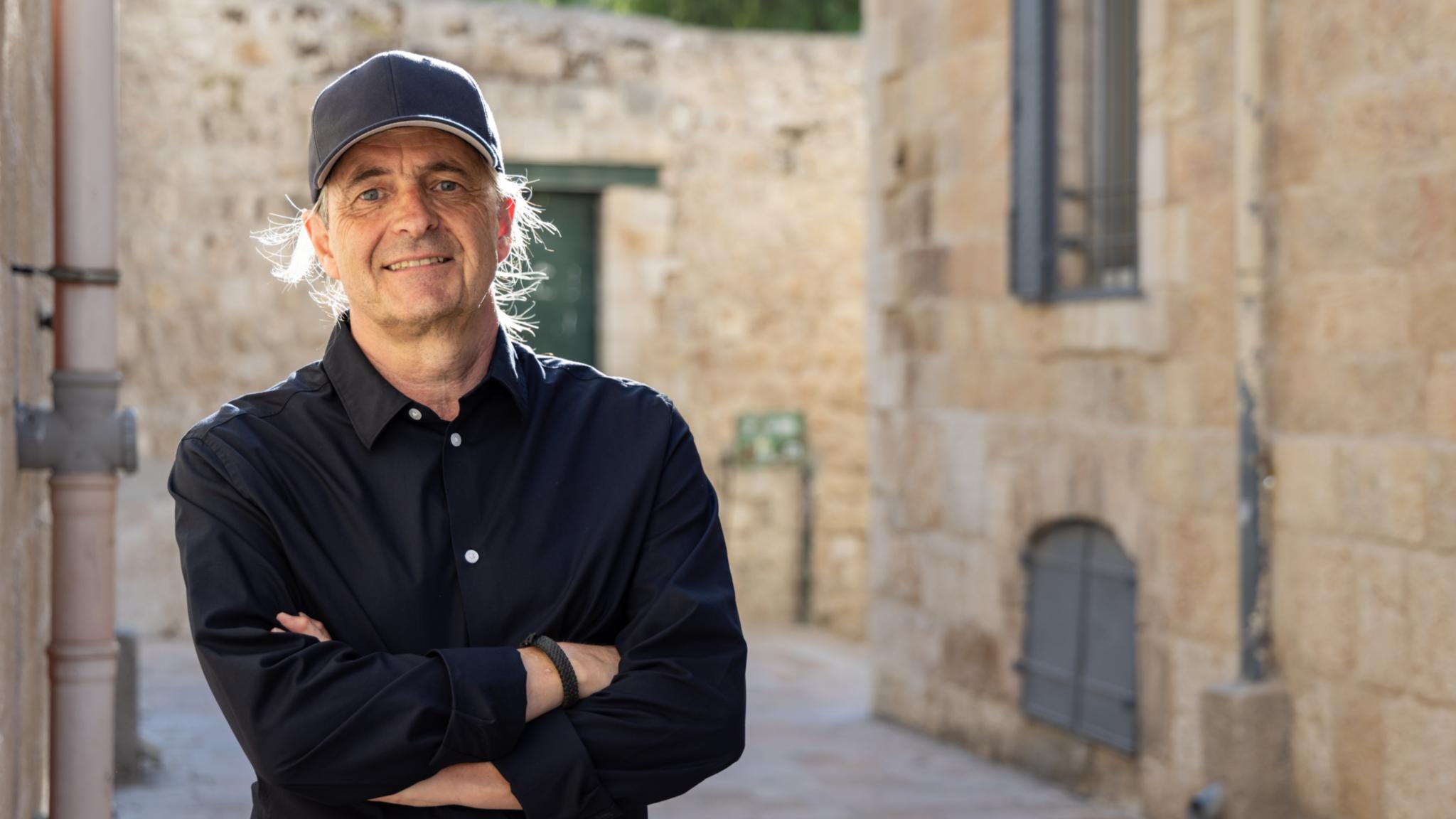 Mann mit schwarzem Hemd und Kappe steht lächelnd in einer Gasse. (KI-Text)