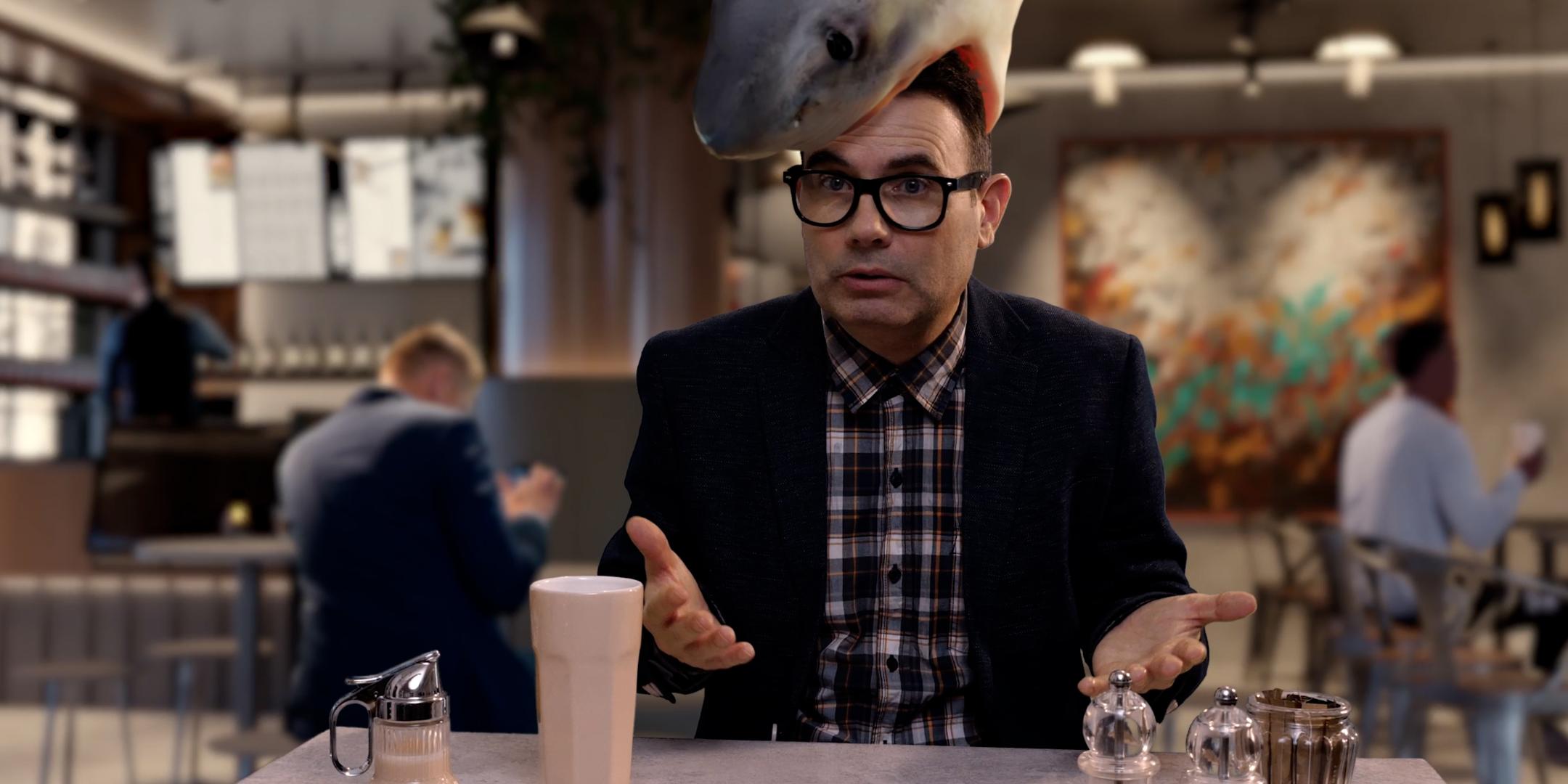 Eine Person sitzt an einem Tisch in einem modern eingerichteten Café oder Restaurant. Auf dem Kopf der Person befindet sich die realistisch dargestellte Figur eines großen Hais, der den Kopf von oben zu umschließen scheint. Vor der Person stehen ein helles Getränk in einem hohen Glas sowie mehrere Gewürzstreuer und eine kleine Karaffe. Im Hintergrund sind weitere Personen an Tischen zu sehen sowie eine Bar und dekorative Wandgestaltung.