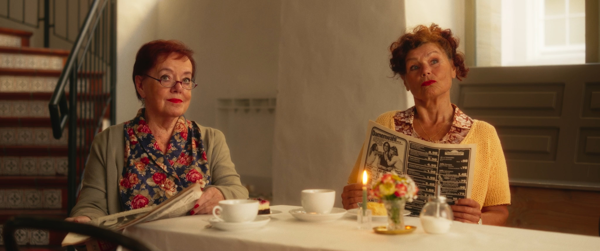 Zwei Personen sitzen an einem gedeckten Tisch in einem hellen Raum. Jede Person hält eine Zeitung in den Händen. Auf dem Tisch stehen zwei weiße Tassen mit Untertassen, eine kleine Vase mit bunten Blumen und eine brennende Kerze auf einem goldenen Teller. Im Hintergrund sind eine Treppe mit verzierten Fliesen, eine helle Wand und eine große Tür zu sehen. Das Licht wirkt warm und ruhig und erinnert an ein Café oder einen Aufenthaltsraum.