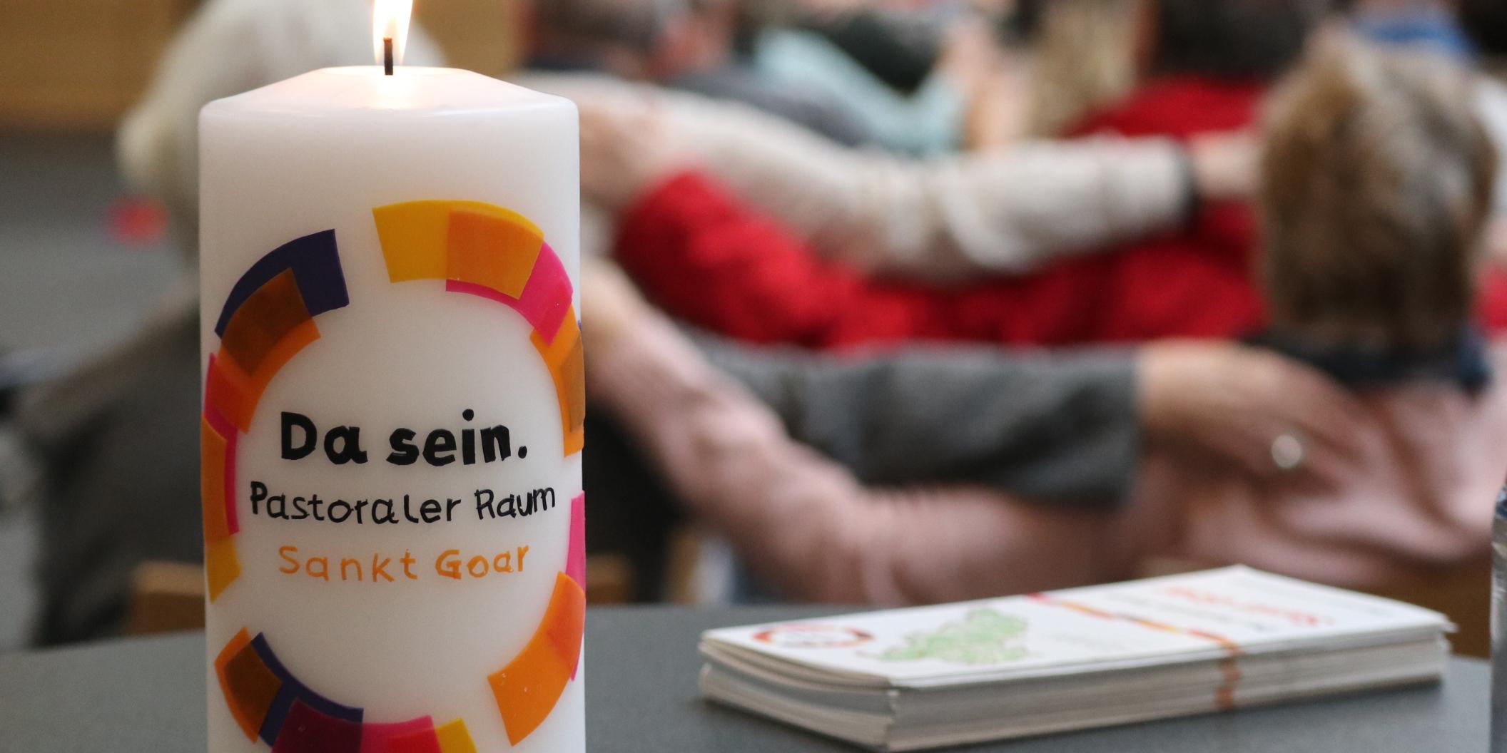 Eine Kerze mit der Aufschrift 'Da sein' steht auf einem Tisch, im Hintergrund sieht man Menschen, die Arme aufeinander legen.