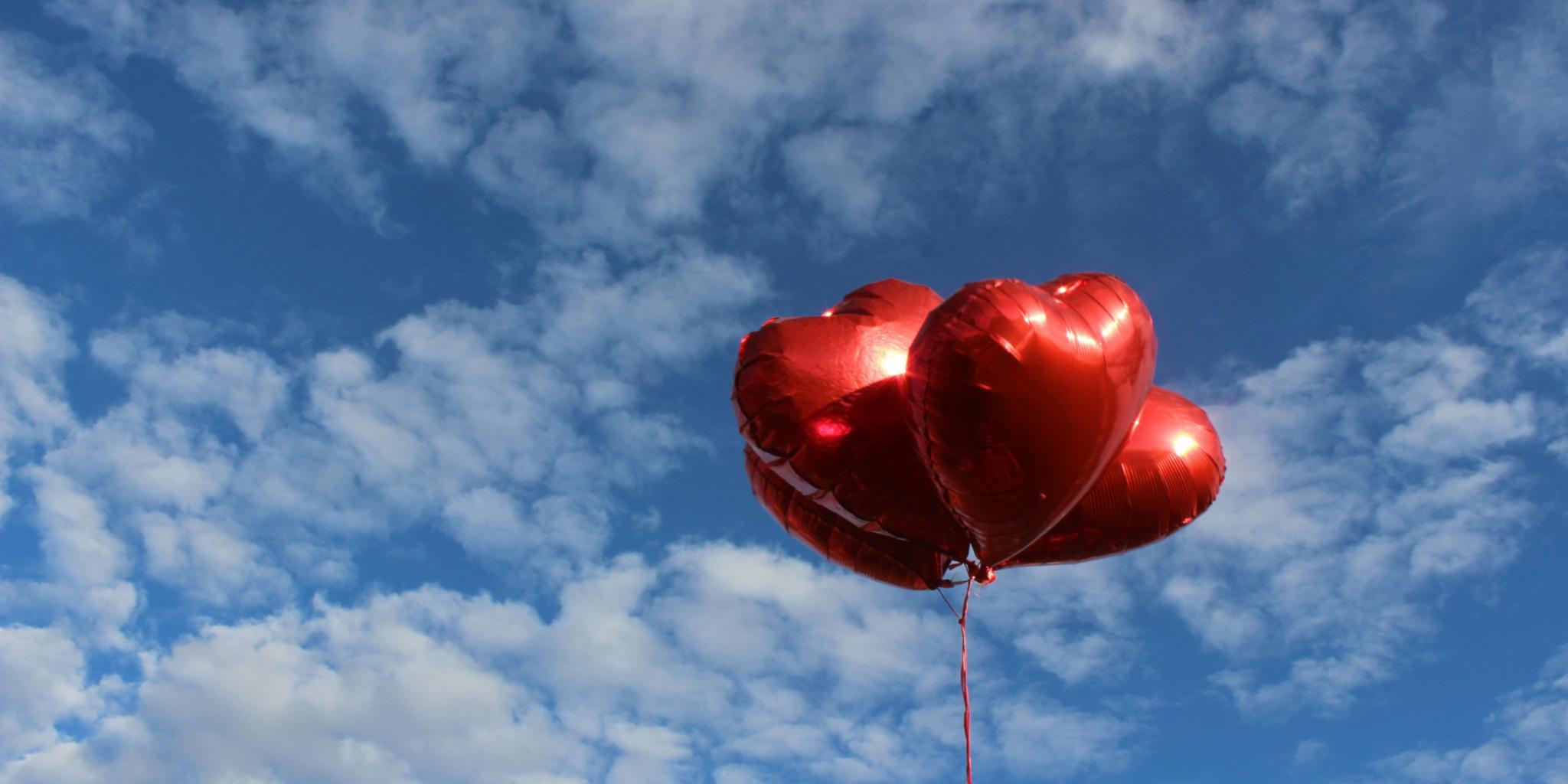 Vor einem blauen Himmel mit verstreuten weißen Wolken schweben drei glänzende, rote, herzförmige Ballons. Sie hängen an dünnen roten Schnüren und heben sich deutlich vom Himmel ab.
