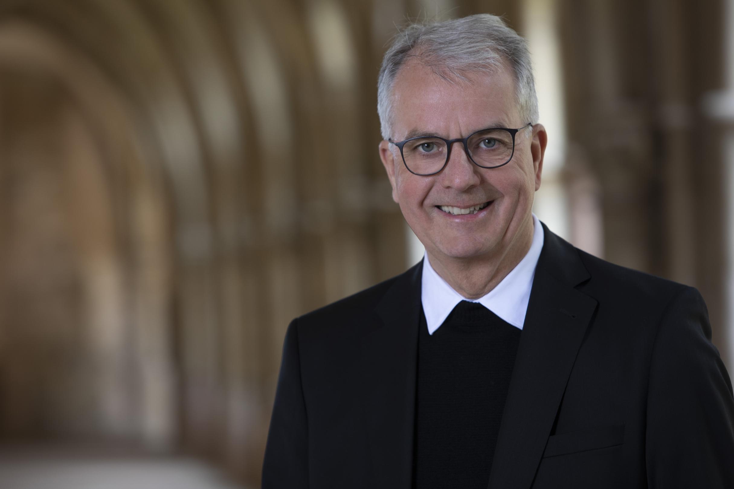 Person im dunklen Anzug in einem Kreuzgang in warmem Licht.