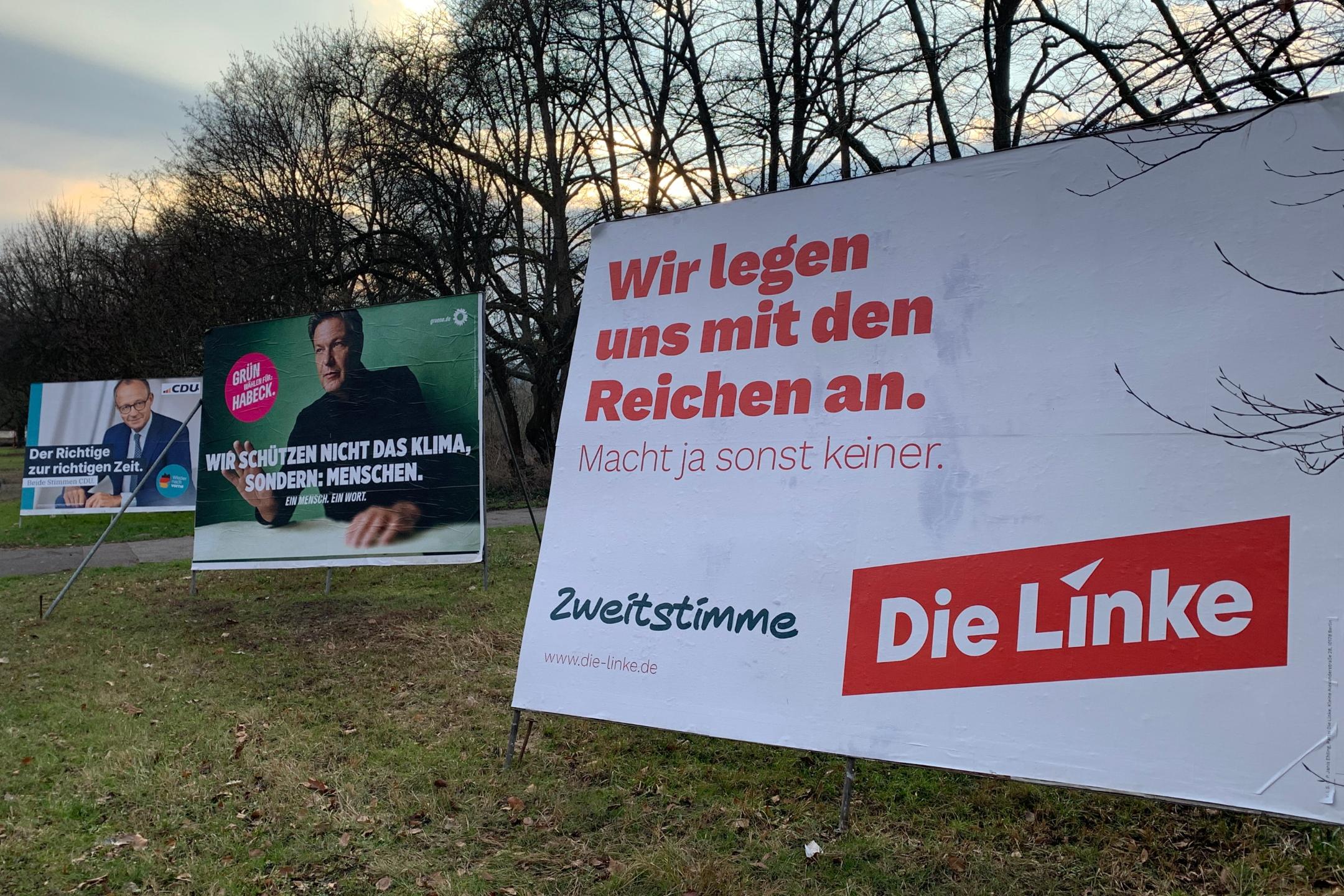 Das Bild zeigt drei große Wahlplakate verschiedener Parteien auf einer Wiese mit kahlen Bäumen im Hintergrund. Im Vordergrund steht ein Plakat der Partei Die Linke mit der Aufschrift „Wir legen uns mit den Reichen an.“ Daneben sind Plakate der Grünen mit Robert Habeck und der CDU mit Friedrich Merz zu sehen, jeweils mit ihren eigenen Wahlbotschaften.  (Alt-Text erstellt mit ChatGPT)