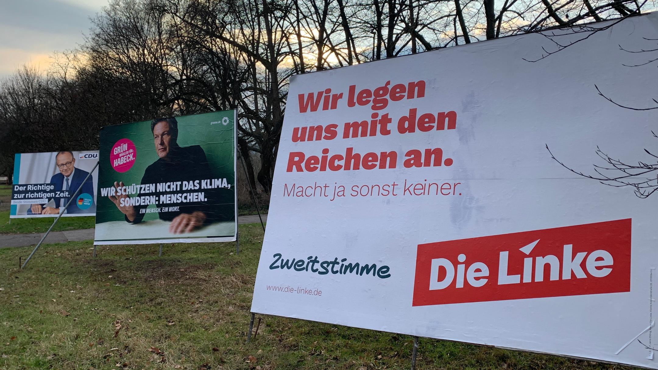 Das Bild zeigt drei große Wahlplakate verschiedener Parteien auf einer Wiese mit kahlen Bäumen im Hintergrund. Im Vordergrund steht ein Plakat der Partei Die Linke mit der Aufschrift „Wir legen uns mit den Reichen an.“ Daneben sind Plakate der Grünen mit Robert Habeck und der CDU mit Friedrich Merz zu sehen, jeweils mit ihren eigenen Wahlbotschaften.  (Alt-Text erstellt mit ChatGPT)