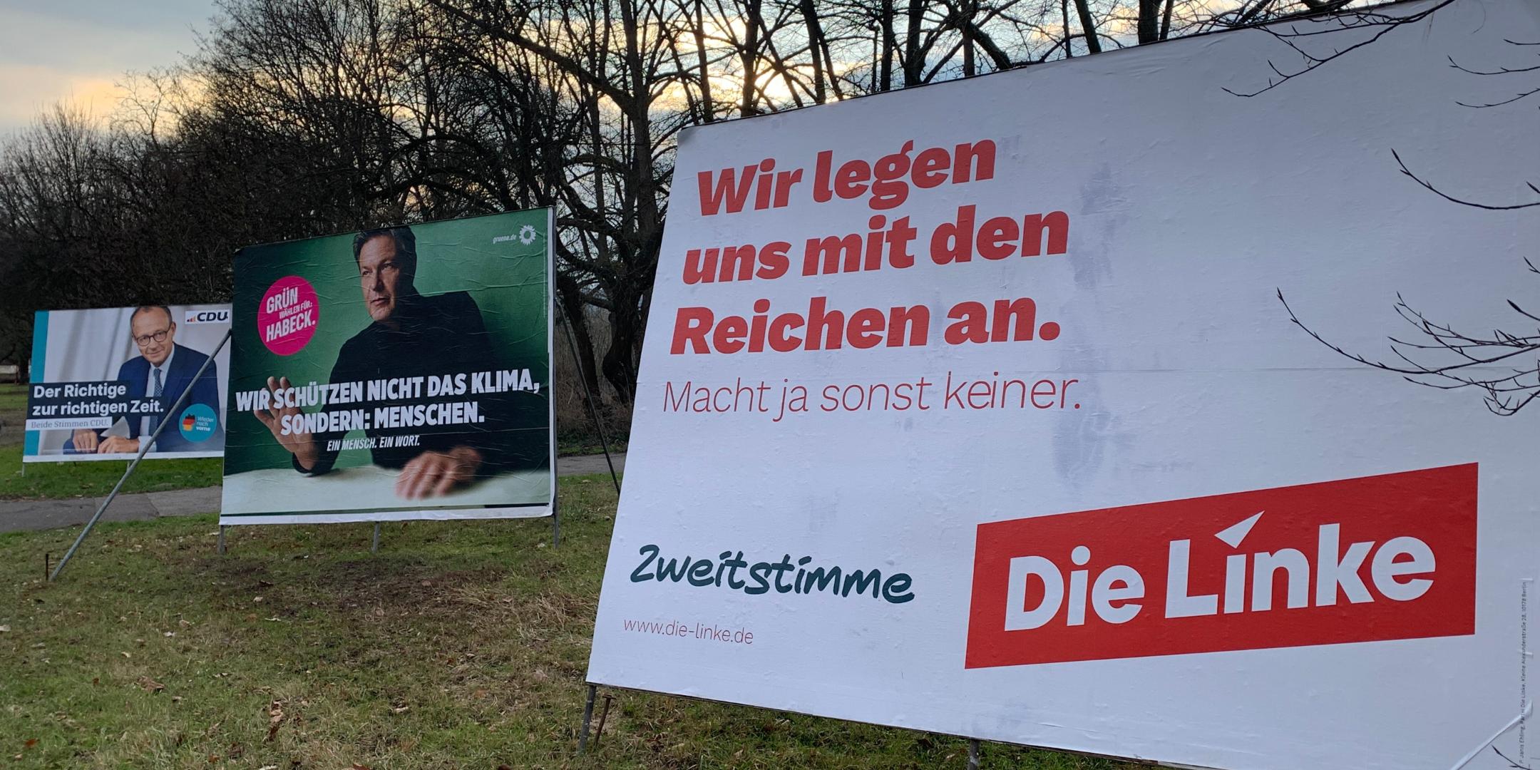 Das Bild zeigt drei große Wahlplakate verschiedener Parteien auf einer Wiese mit kahlen Bäumen im Hintergrund. Im Vordergrund steht ein Plakat der Partei Die Linke mit der Aufschrift „Wir legen uns mit den Reichen an.“ Daneben sind Plakate der Grünen mit Robert Habeck und der CDU mit Friedrich Merz zu sehen, jeweils mit ihren eigenen Wahlbotschaften.  (Alt-Text erstellt mit ChatGPT)