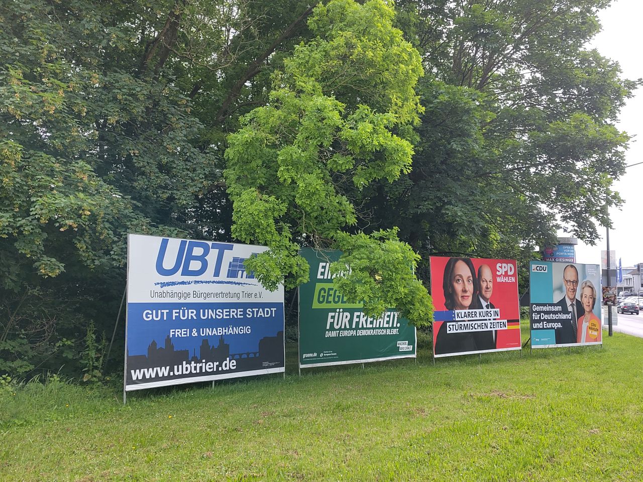 Das Foto zeigt vier Plakatwände mit Wahlplakaten zur Kommunal- und Europawahl, die nebeneinander auf einer Wiese am Rand einer Straße stehen; dahinter Bäume. Die Plakate (von links nach rechts) werben für die Unabhängige Bürgervertretung Trier (UBT), die Grünen, die SPD und die CDU.