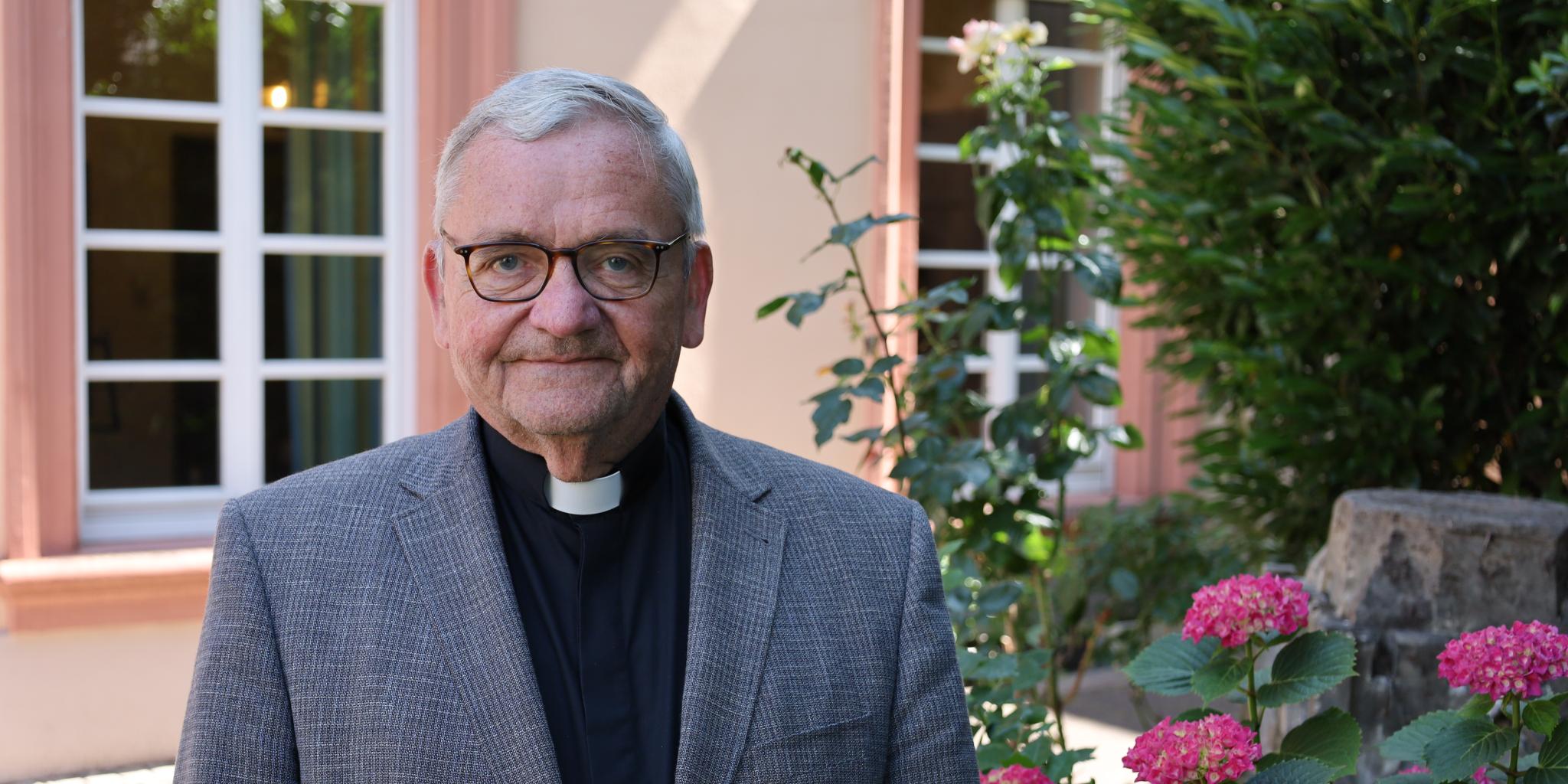 Ein Pfarrer steht vor einem Gebäude. Neben ihm ist ein blühender Hortensienbusch