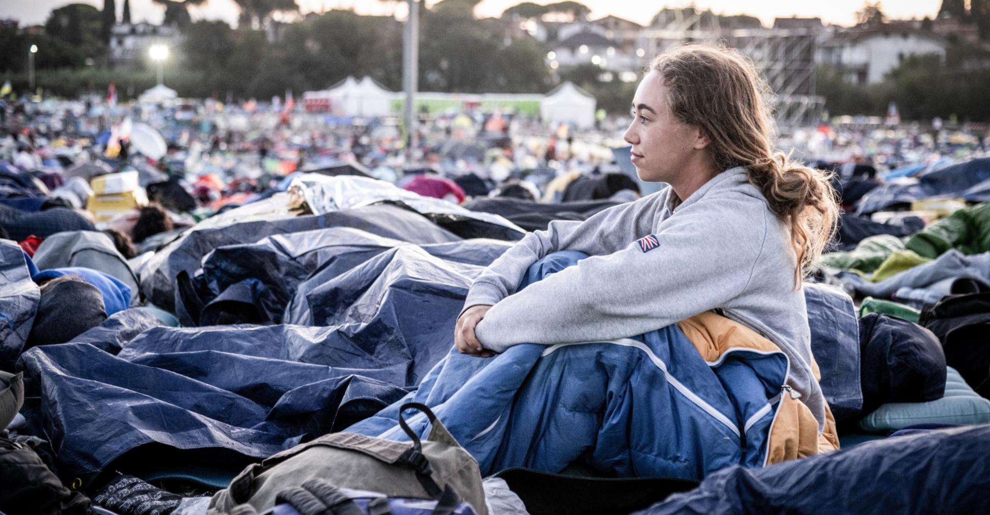 Am Morgen nach der Gebetsnacht beim Weltjugendtreffen 2025 in Rom: Eine junge Frau sitzt in ihrem Schlafsack und blickt über das Gelände.