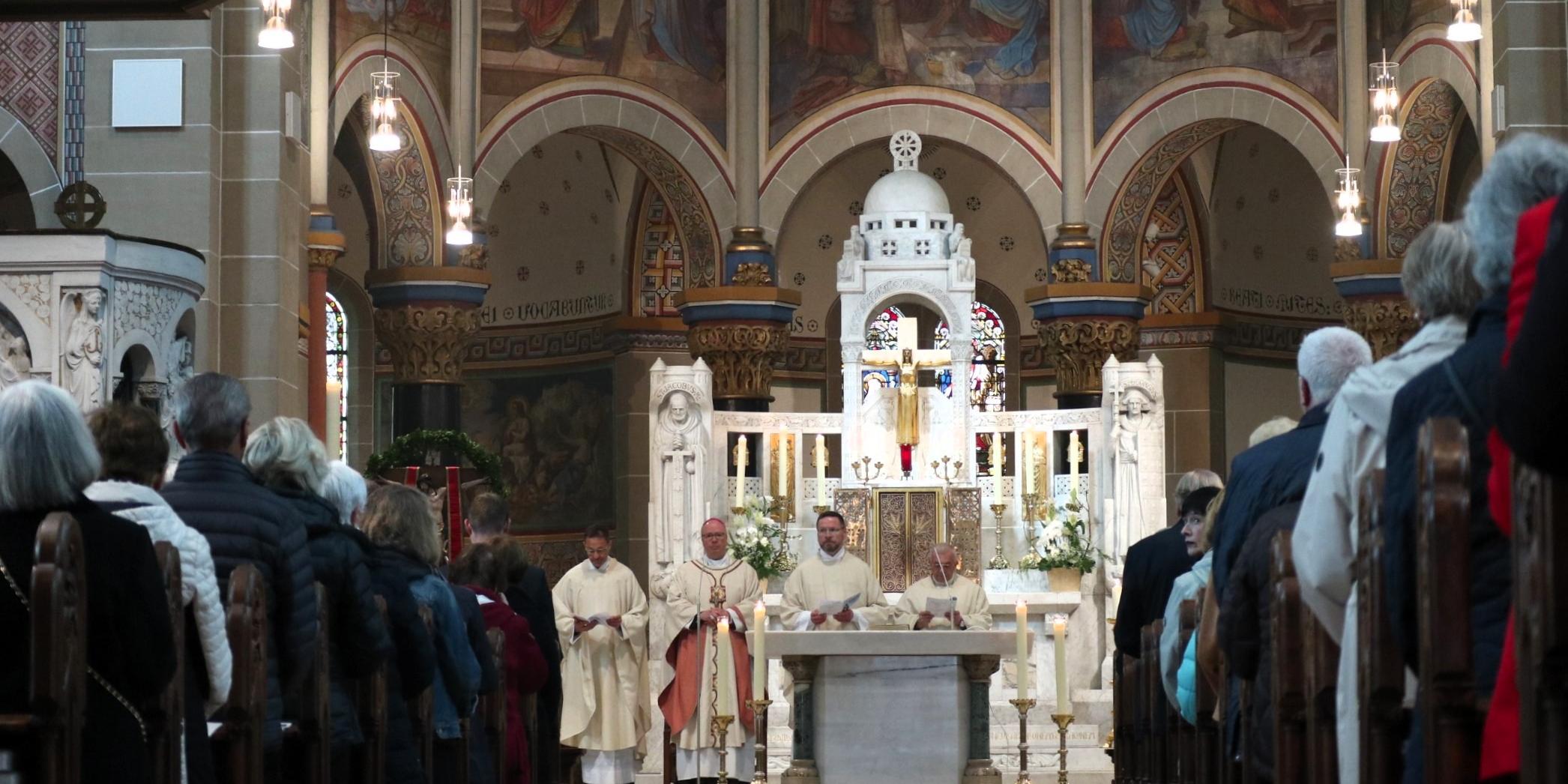 Priester stehen an einem weißen Altar, zu sehen ist auch ein langer Mittelgang mit vielen Personen, die in Kirchenbänken sitzen.