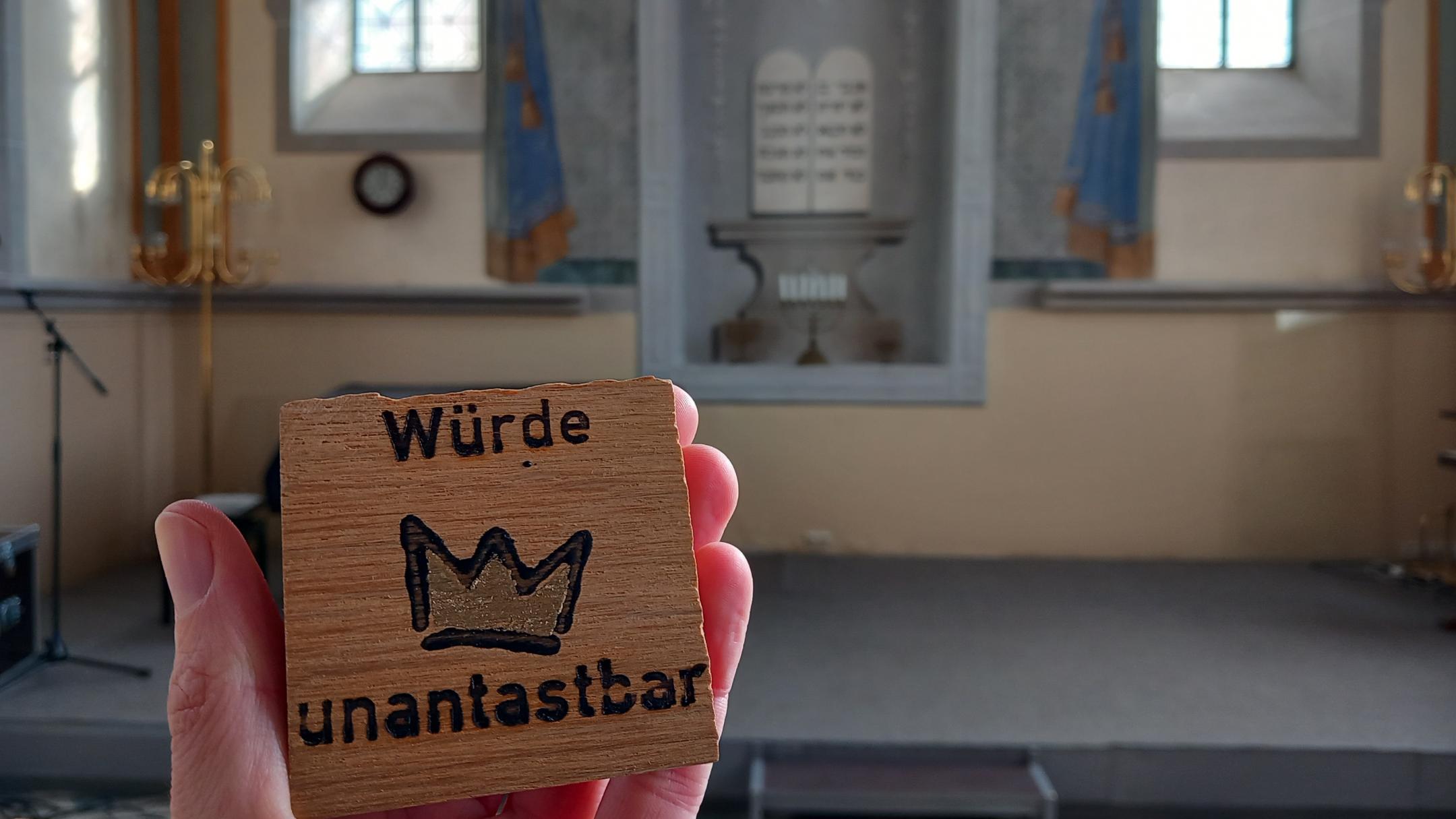 Ein Mensch hält ein kleines Holzschild in der Hand, auf dem „Würde unantastbar“ mit einer gezeichneten Krone dazwischen steht. Im Hintergrund ist der Innenraum einer Synagoge zu sehen, mit einer Bühne, goldenen Leuchtern und zwei hohen Fenstern. An der Wand hinter der Bühne ist ein gemaltes Bild mit zwei Gesetzestafeln in hebräischer Schrift zu erkennen. (Alt-Text erstellt mit Unterstützung von ChatGPT)