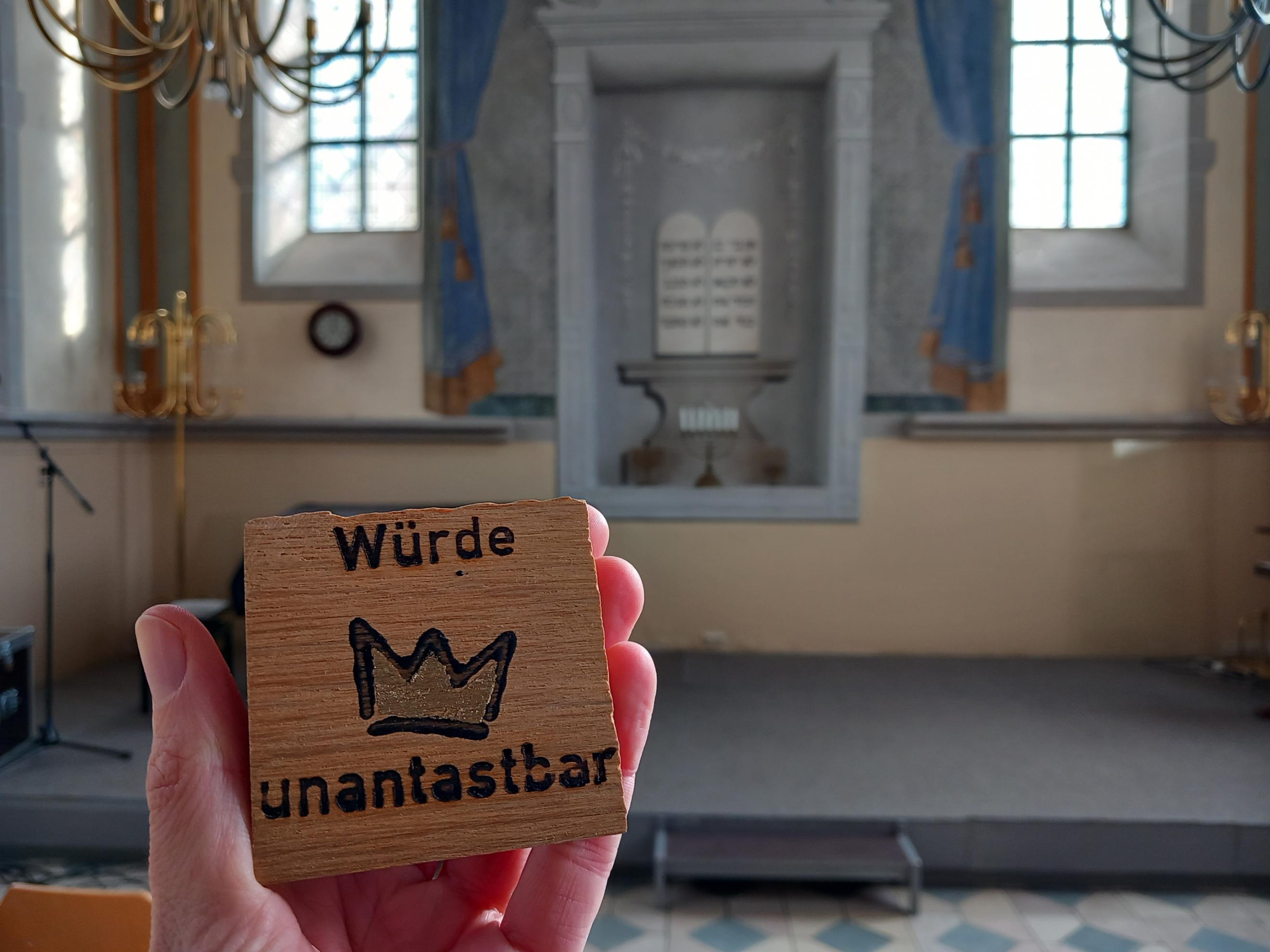 Ein Mensch hält ein kleines Holzschild in der Hand, auf dem „Würde unantastbar“ mit einer gezeichneten Krone dazwischen steht. Im Hintergrund ist der Innenraum einer Synagoge zu sehen, mit einer Bühne, goldenen Leuchtern und zwei hohen Fenstern. An der Wand hinter der Bühne ist ein gemaltes Bild mit zwei Gesetzestafeln in hebräischer Schrift zu erkennen. (Alt-Text erstellt mit Unterstützung von ChatGPT)
