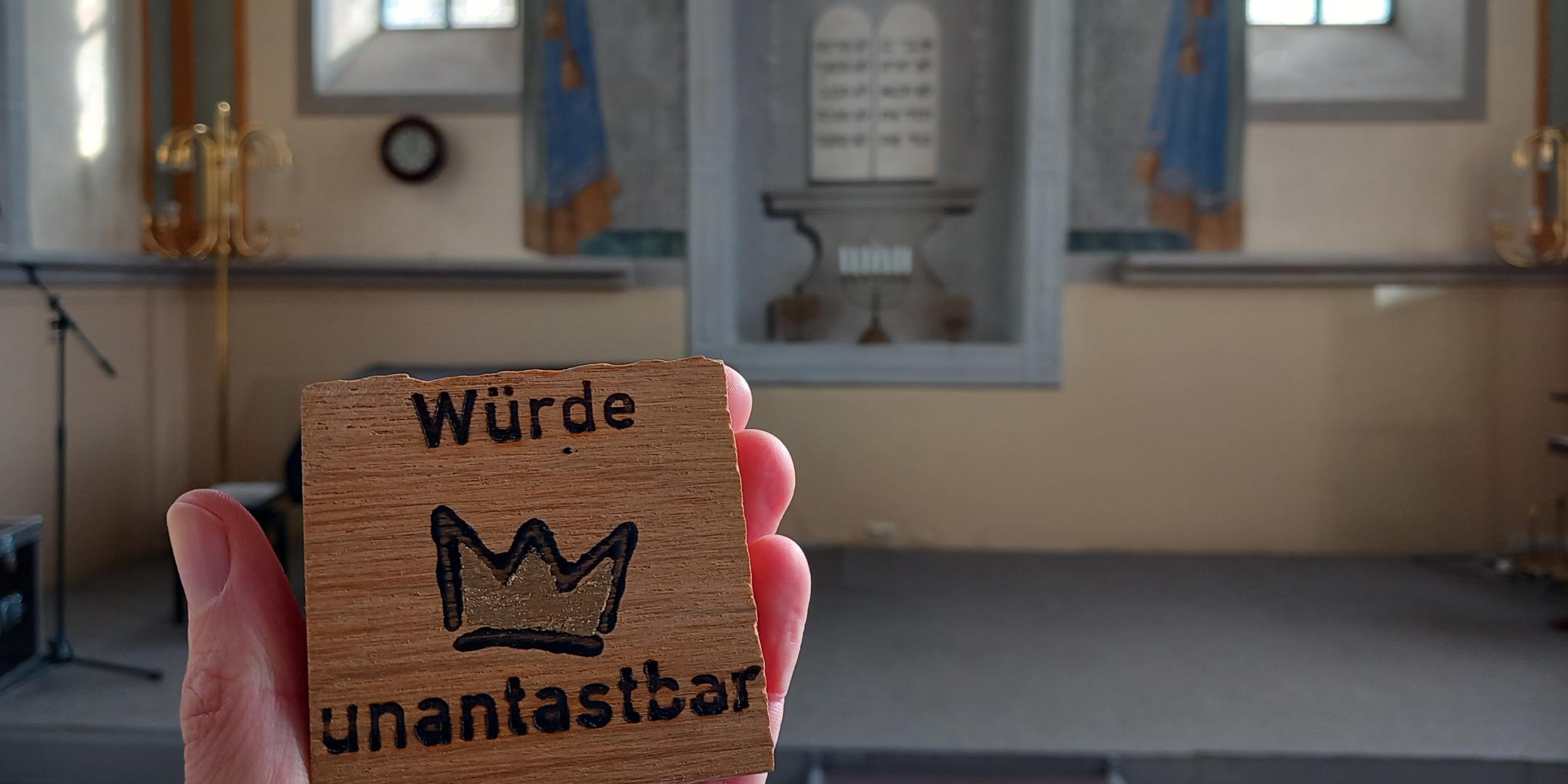 Ein Mensch hält ein kleines Holzschild in der Hand, auf dem „Würde unantastbar“ mit einer gezeichneten Krone dazwischen steht. Im Hintergrund ist der Innenraum einer Synagoge zu sehen, mit einer Bühne, goldenen Leuchtern und zwei hohen Fenstern. An der Wand hinter der Bühne ist ein gemaltes Bild mit zwei Gesetzestafeln in hebräischer Schrift zu erkennen. (Alt-Text erstellt mit Unterstützung von ChatGPT)