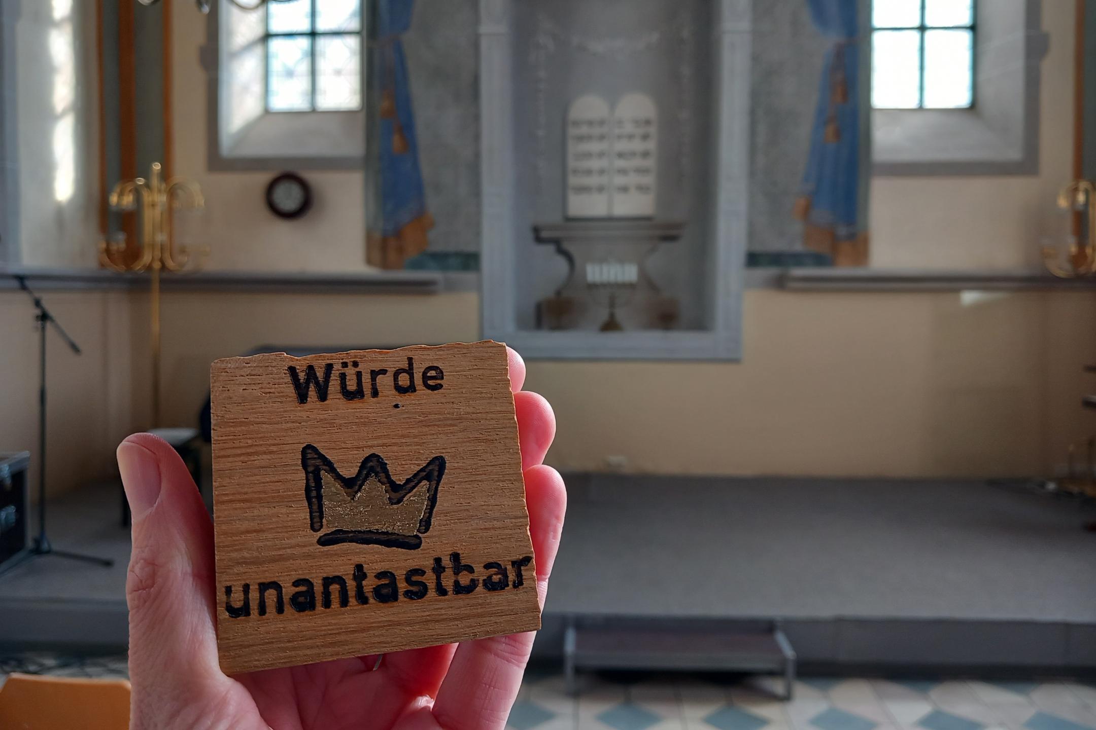 Ein Mensch hält ein kleines Holzschild in der Hand, auf dem „Würde unantastbar“ mit einer gezeichneten Krone dazwischen steht. Im Hintergrund ist der Innenraum einer Synagoge zu sehen, mit einer Bühne, goldenen Leuchtern und zwei hohen Fenstern. An der Wand hinter der Bühne ist ein gemaltes Bild mit zwei Gesetzestafeln in hebräischer Schrift zu erkennen. (Alt-Text erstellt mit Unterstützung von ChatGPT)
