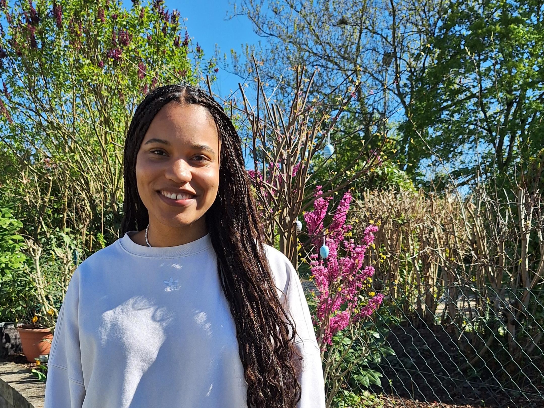 Eine junge Frau mit langen, geflochtenen Haaren – es handelt sich um die Kugelstoßerin Yemisi Ogunleye - steht lächelnd in einem sonnigen Garten vor einem Beet mit bunten Frühlingsblumen, darunter Tulpen und Hyazinthen. Sie trägt ein helles Sweatshirt und eine weiße Hose. Im Hintergrund sind blühende Sträucher, Bäume und ein blauer Himmel zu sehen.