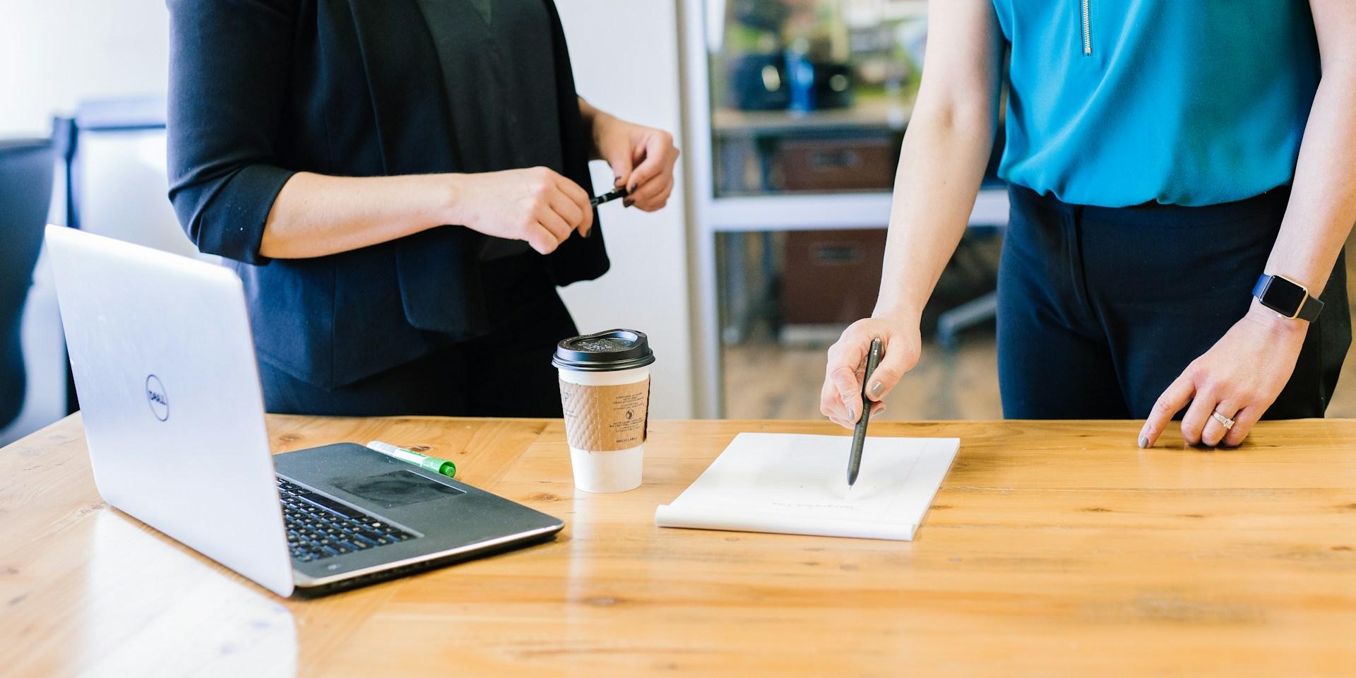 Das Foto zeigt zwei Frauen in einem Büro. Auf dem Tisch liegen ein Laptop, ein Kaffeebecher und ein grüner Marker. Im Hintergrund sind Glaswände und Büromöbel zu sehen.