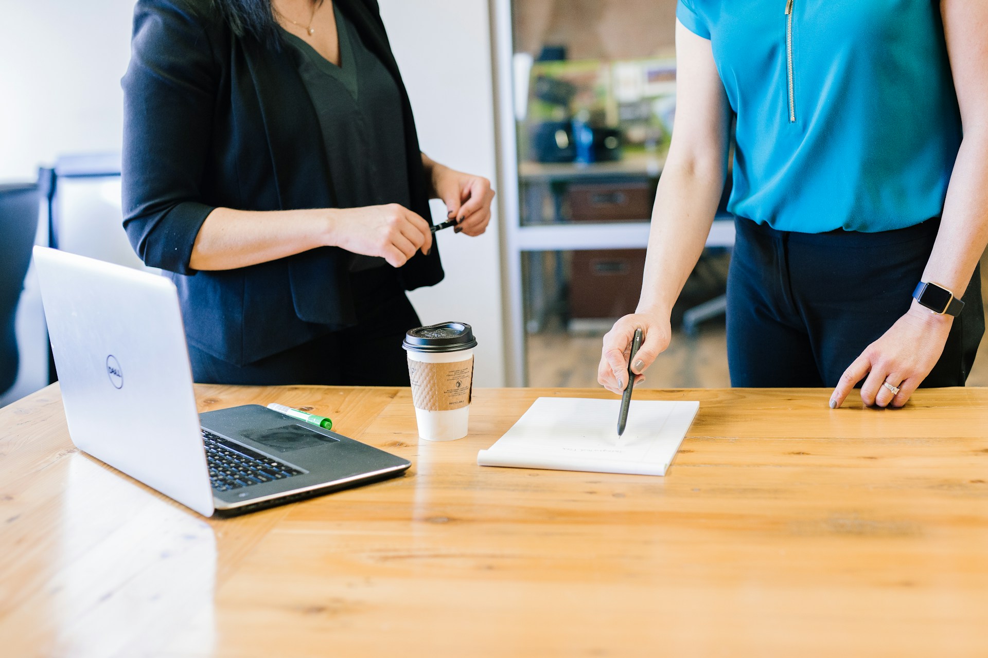 Das Foto zeigt zwei Frauen in einem Büro. Auf dem Tisch liegen ein Laptop, ein Kaffeebecher und ein grüner Marker. Im Hintergrund sind Glaswände und Büromöbel zu sehen.