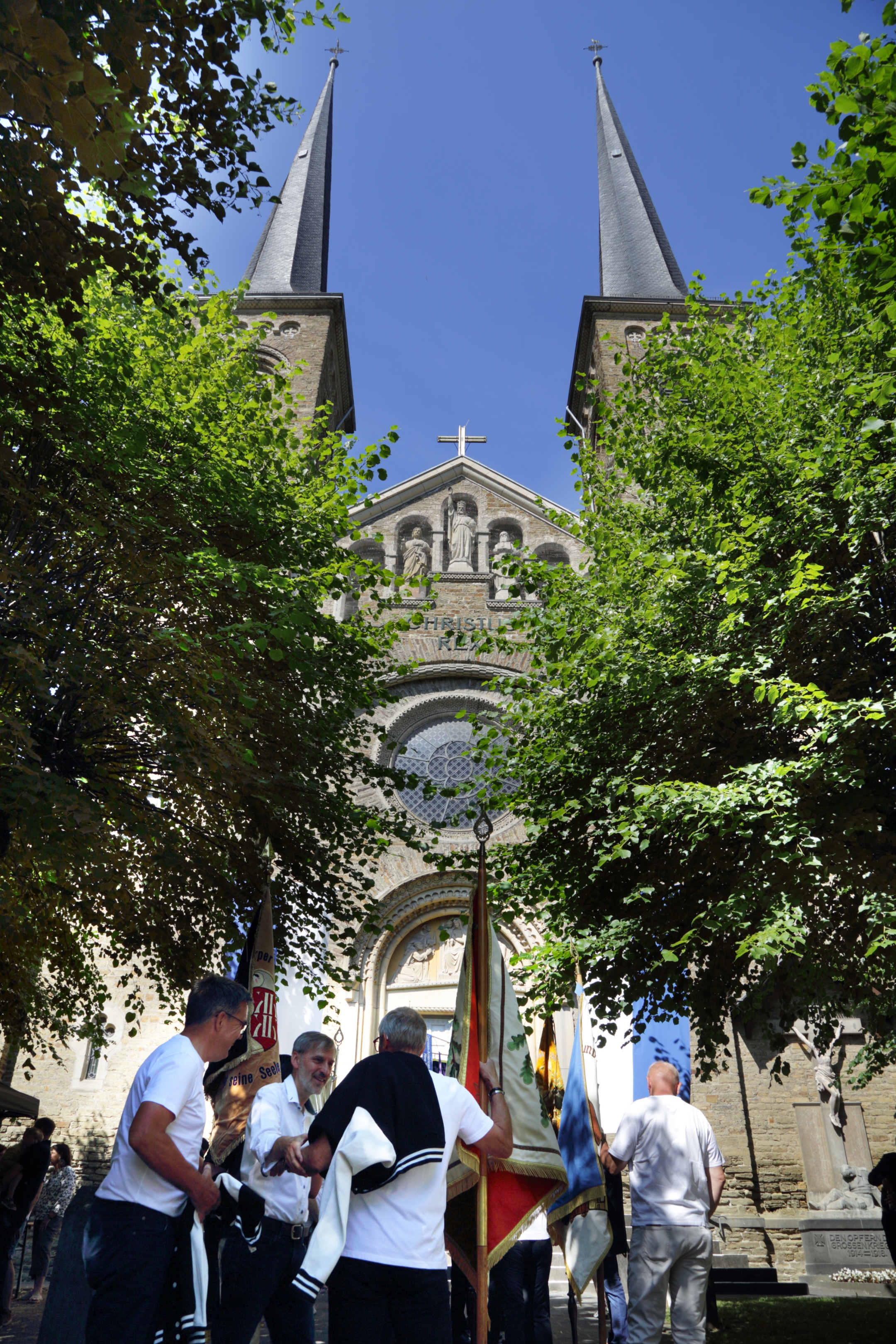 Vor einer Kirche mit Bäumen stehen einige Menschen mit den Fahnen der Ortsvereine