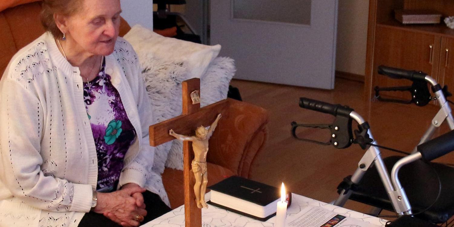 Eine ältere Person sitzt in einem Wohnzimmer an einem Tisch, der mit einer weißen, gemusterten Tischdecke bedeckt ist. Auf dem Tisch stehen ein hölzernes Kruzifix, zwei brennende Kerzen, eine Bibel und ein kleiner Zweig in einer Glasschale. Neben dem Tisch befindet sich ein Rollator. In der Umgebung sind ein Sofa mit Kissen und ein Regal zu sehen.