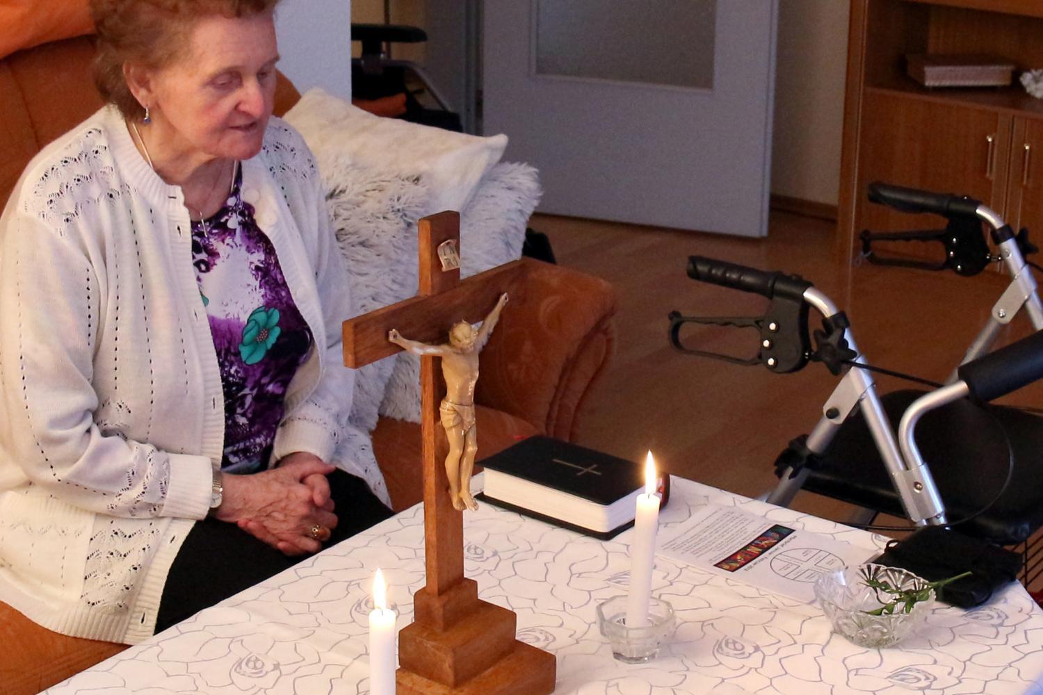 Eine ältere Person sitzt in einem Wohnzimmer an einem Tisch, der mit einer weißen, gemusterten Tischdecke bedeckt ist. Auf dem Tisch stehen ein hölzernes Kruzifix, zwei brennende Kerzen, eine Bibel und ein kleiner Zweig in einer Glasschale. Neben dem Tisch befindet sich ein Rollator. In der Umgebung sind ein Sofa mit Kissen und ein Regal zu sehen.