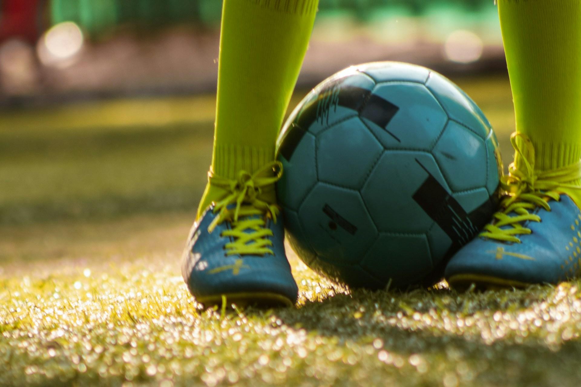 Das Foto zeigt auf einer Wiese in Nahaufnahme zwei Kinderfüße in blauen Turnschuhen und gelb-grünen Strümpfen, die einen Fußball festhalten.