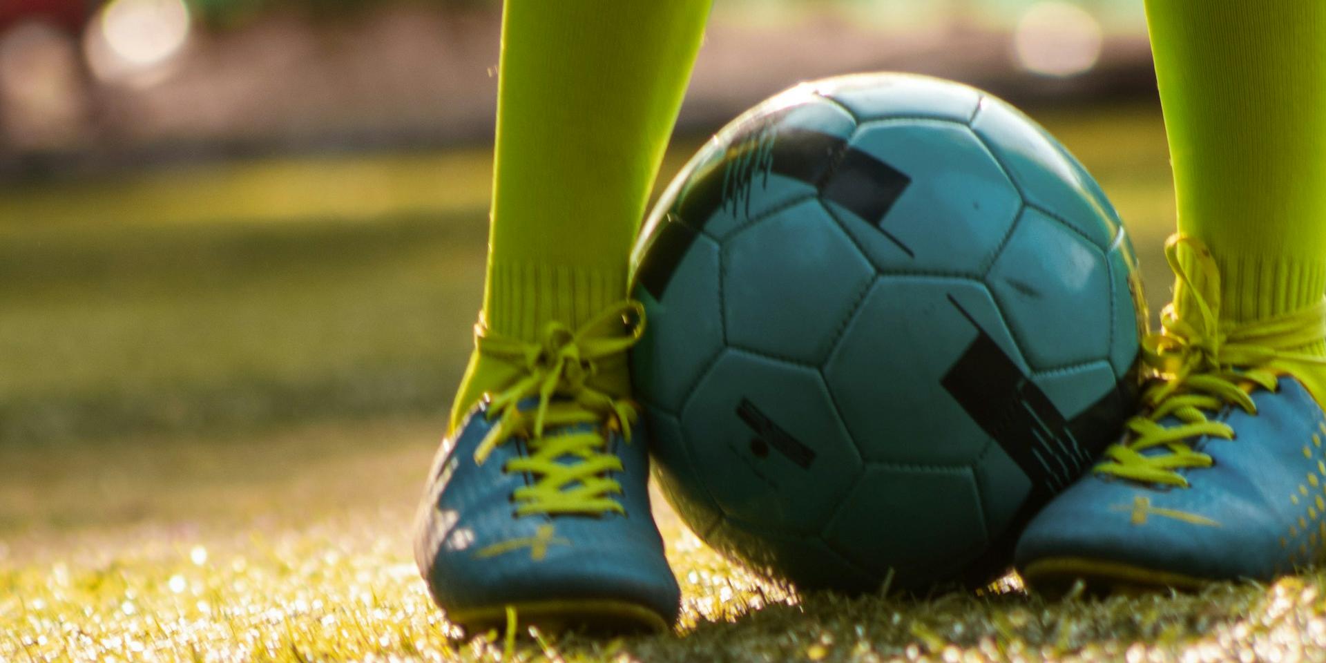 Das Foto zeigt auf einer Wiese in Nahaufnahme zwei Kinderfüße in blauen Turnschuhen und gelb-grünen Strümpfen, die einen Fußball festhalten.