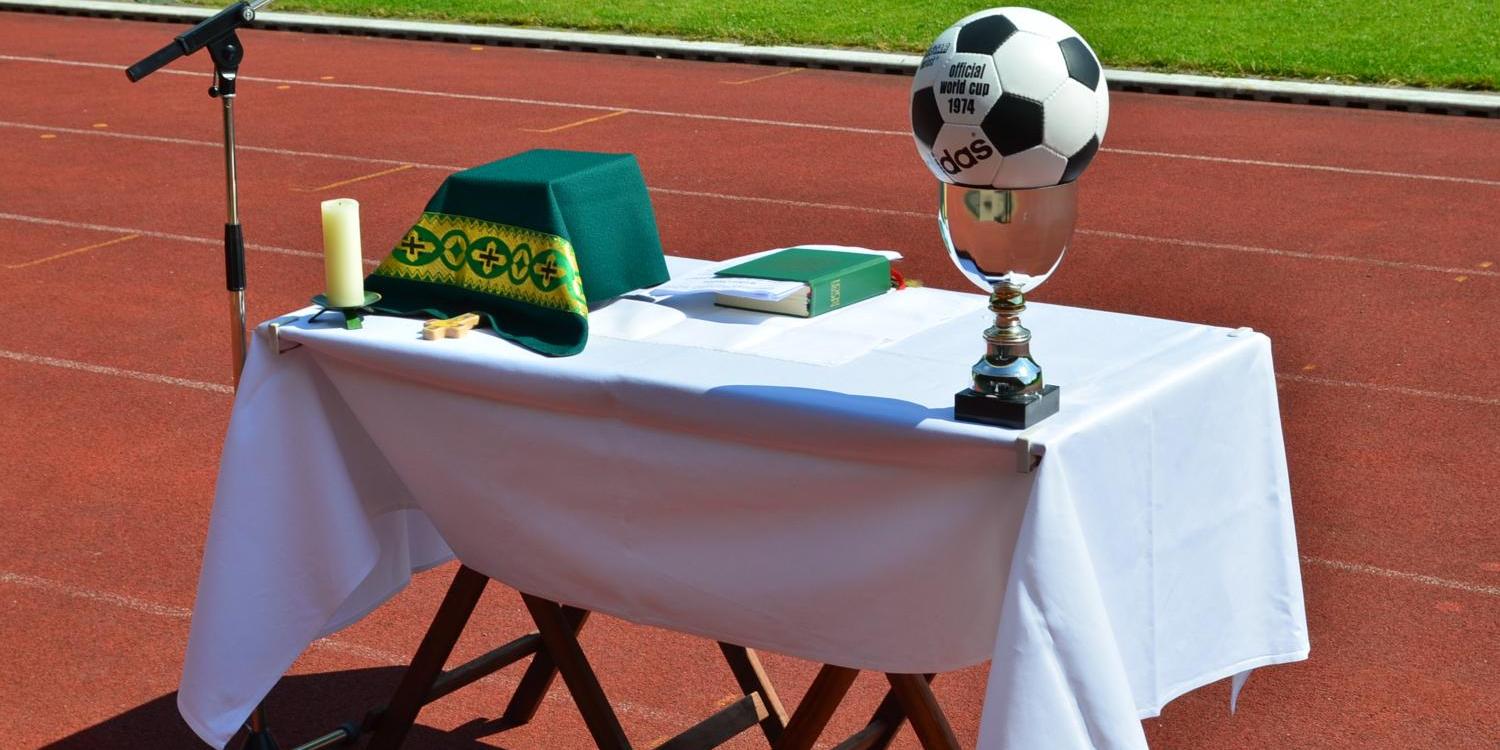 Symbolfoto: Fußball-Gottesdienst Vor dem grünen Rasen eines Stadions ist auf der roten Laufbahn ein provisorischer Altar aufgebaut. Auf dem weiß gedeckten Tisch liegen ein grünes liturgisches Tuch, eine Kerze, ein kleines Holzkreuz und ein liturgisches Buch. Links steht ein Mikrofonständer für eine Freiluftandacht oder einen Gottesdienst. Rechts auf dem Tisch befindet sich ein silberner Pokal, auf dem ein Fußball mit der Aufschrift „Official World Cup 1974“ platziert ist. Die Szene verbindet christliche Symbole mit dem Umfeld eines Sportplatzes.