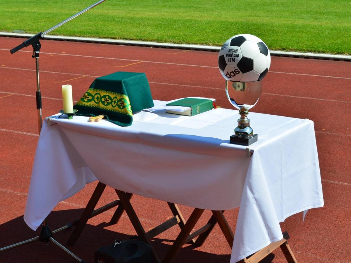 Vor dem grünen Rasen eines Stadions ist auf der roten Laufbahn ein provisorischer Altar aufgebaut. Auf dem weiß gedeckten Tisch liegen ein grünes liturgisches Tuch, eine Kerze, ein kleines Holzkreuz und ein liturgisches Buch. Links steht ein Mikrofonständer für eine Freiluftandacht oder einen Gottesdienst. Rechts auf dem Tisch befindet sich ein silberner Pokal, auf dem ein Fußball mit der Aufschrift „Official World Cup 1974“ platziert ist. Die Szene verbindet christliche Symbole mit dem Umfeld eines Sportplatzes.