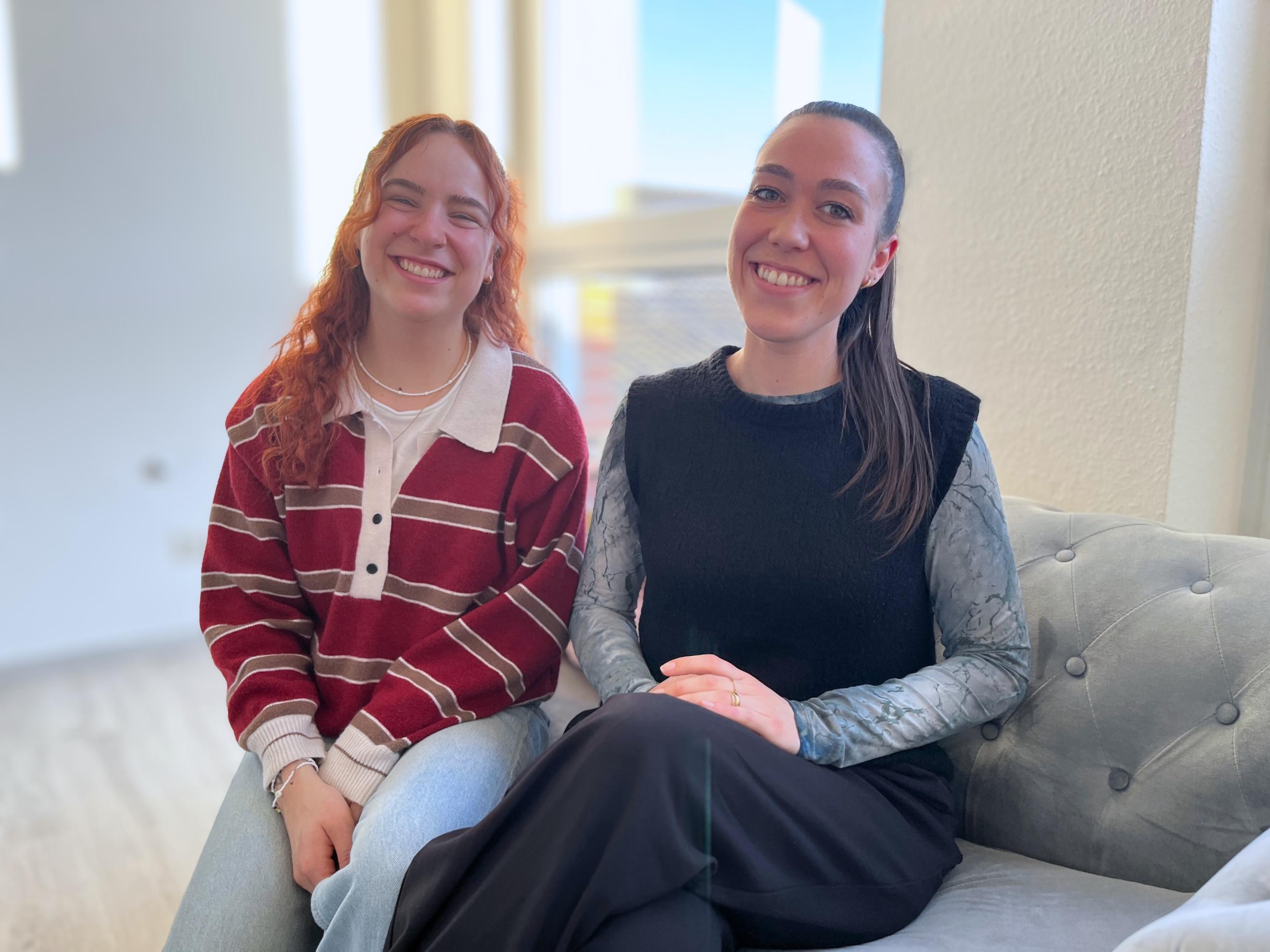 Zwei junge Frauen sitzen nebeneinander auf einem Sofa und lachen in die Kamera.