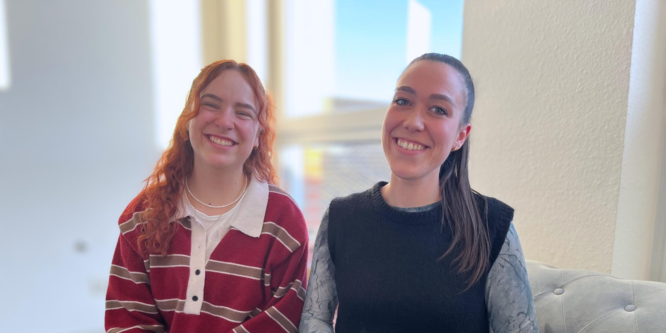 Zwei junge Frauen sitzen nebeneinander auf einem Sofa und lachen in die Kamera.