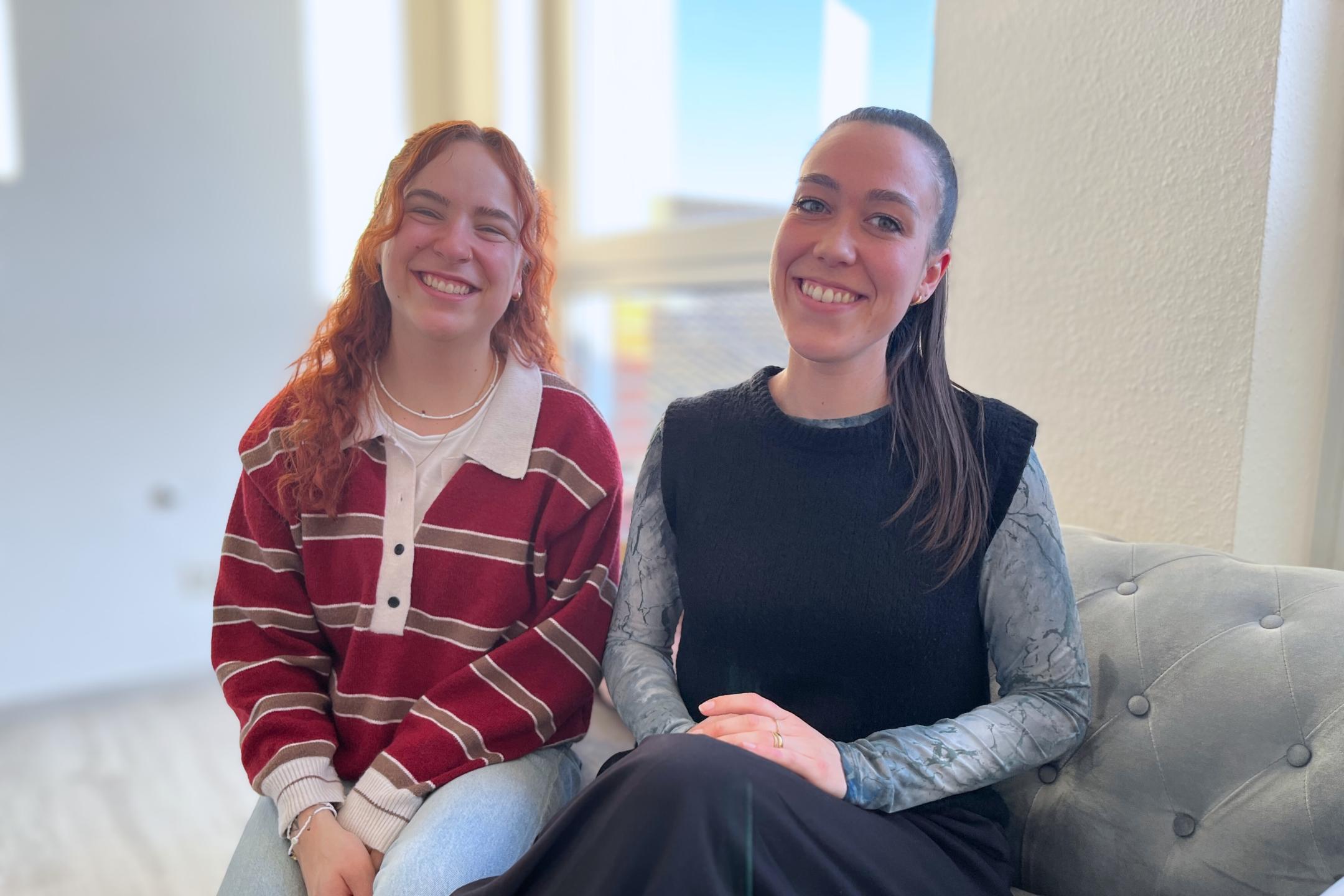 Zwei junge Frauen sitzen nebeneinander auf einem Sofa und lachen in die Kamera.