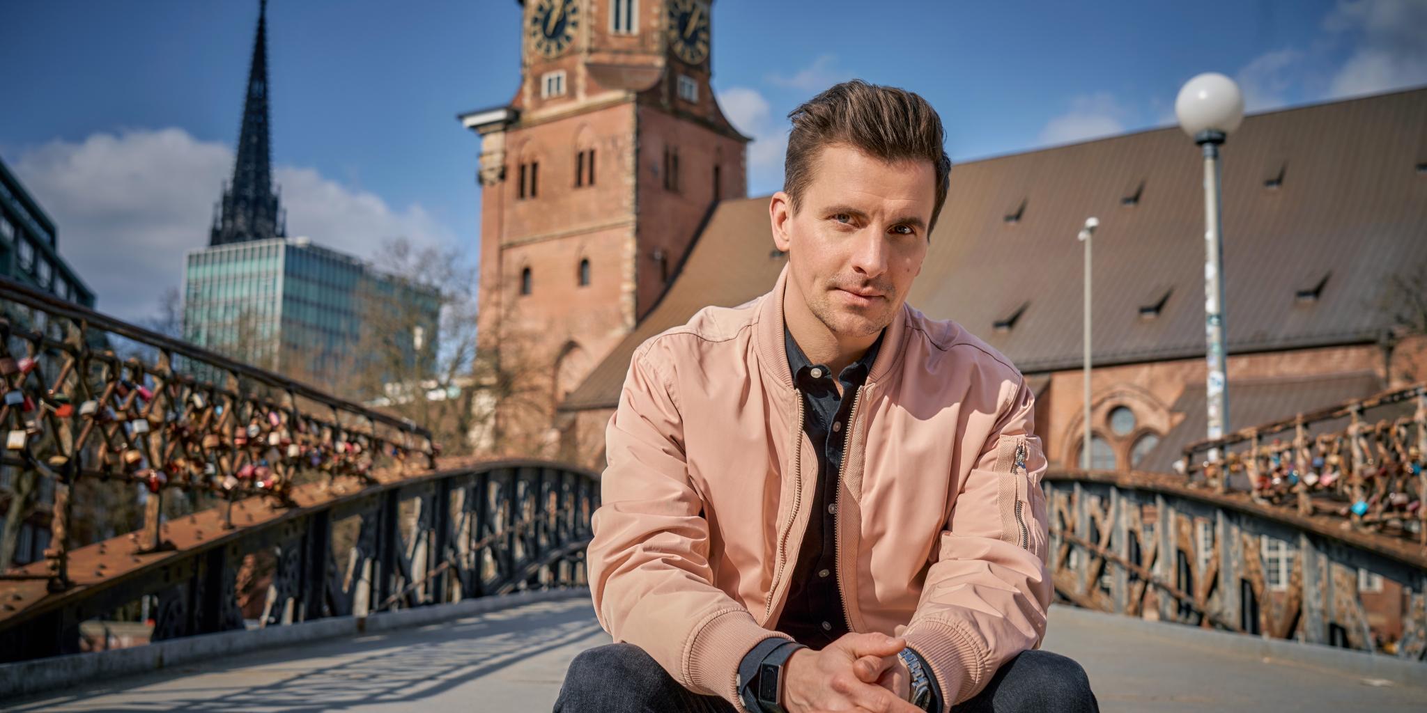 Mann sitzt auf einer Brücke mit Liebesschlössern, Kirche im Hintergrund.