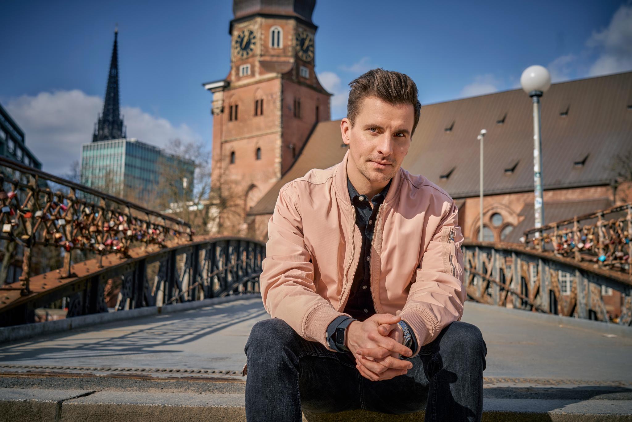 Mann sitzt auf einer Brücke mit Liebesschlössern, Kirche im Hintergrund.