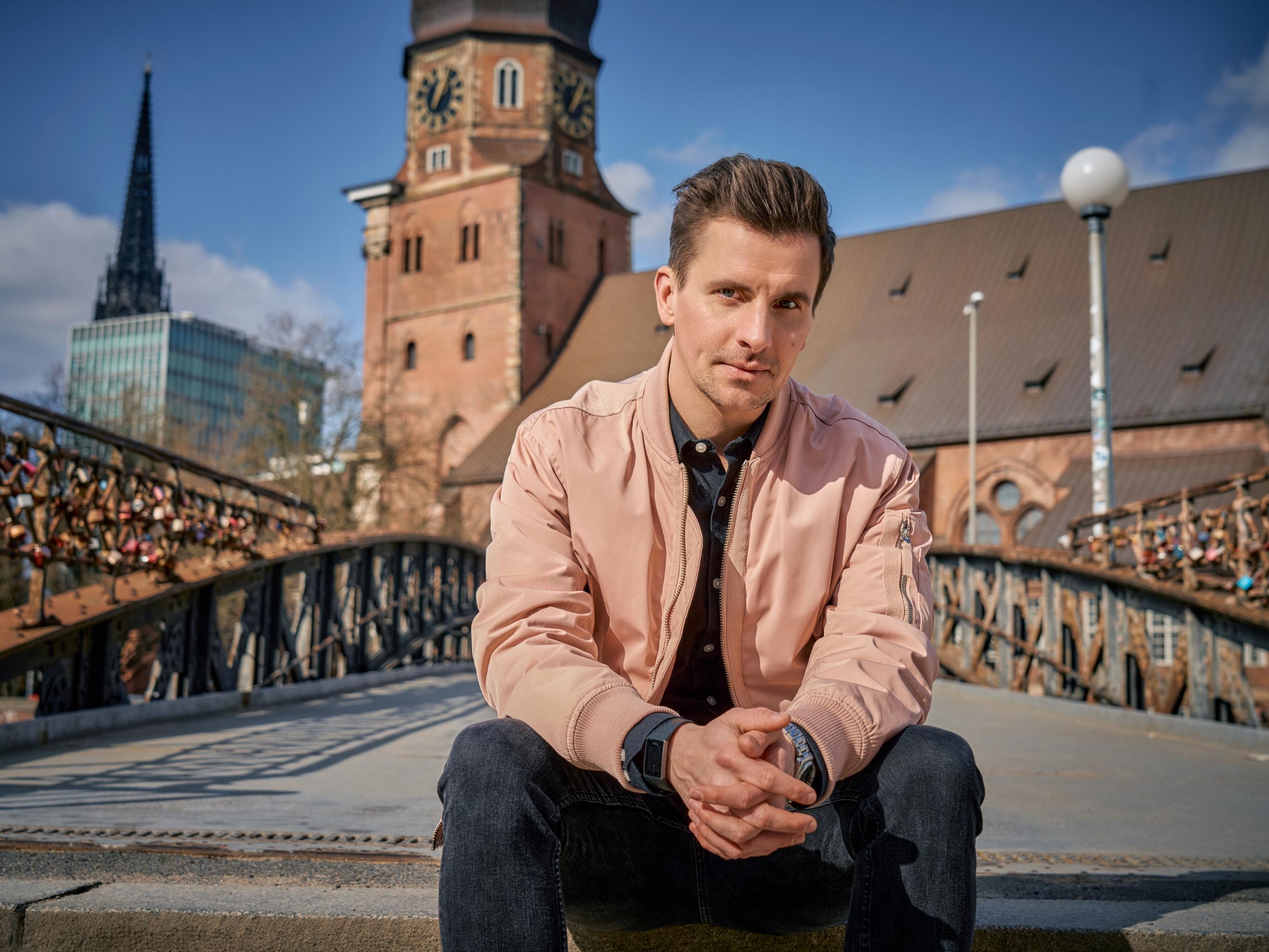Mann sitzt auf einer Brücke mit Liebesschlössern, Kirche im Hintergrund.