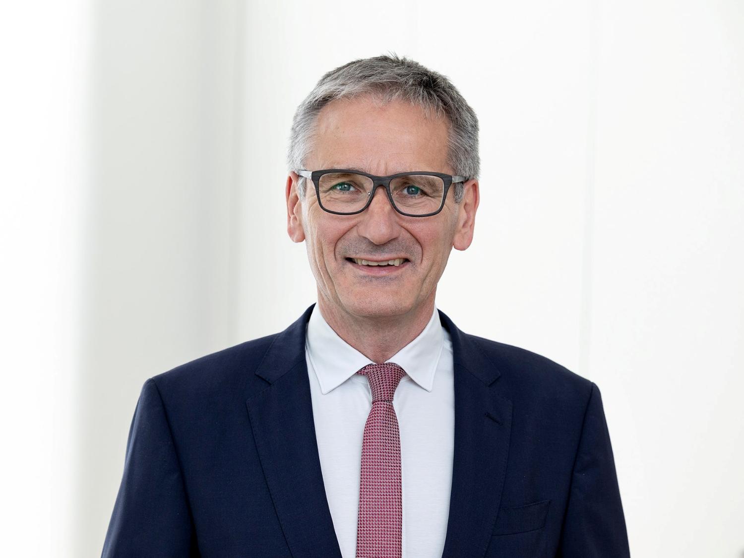 Mann in dunklem Anzug vor hellem Hintergrund blickt geradeaus in die Kamera.