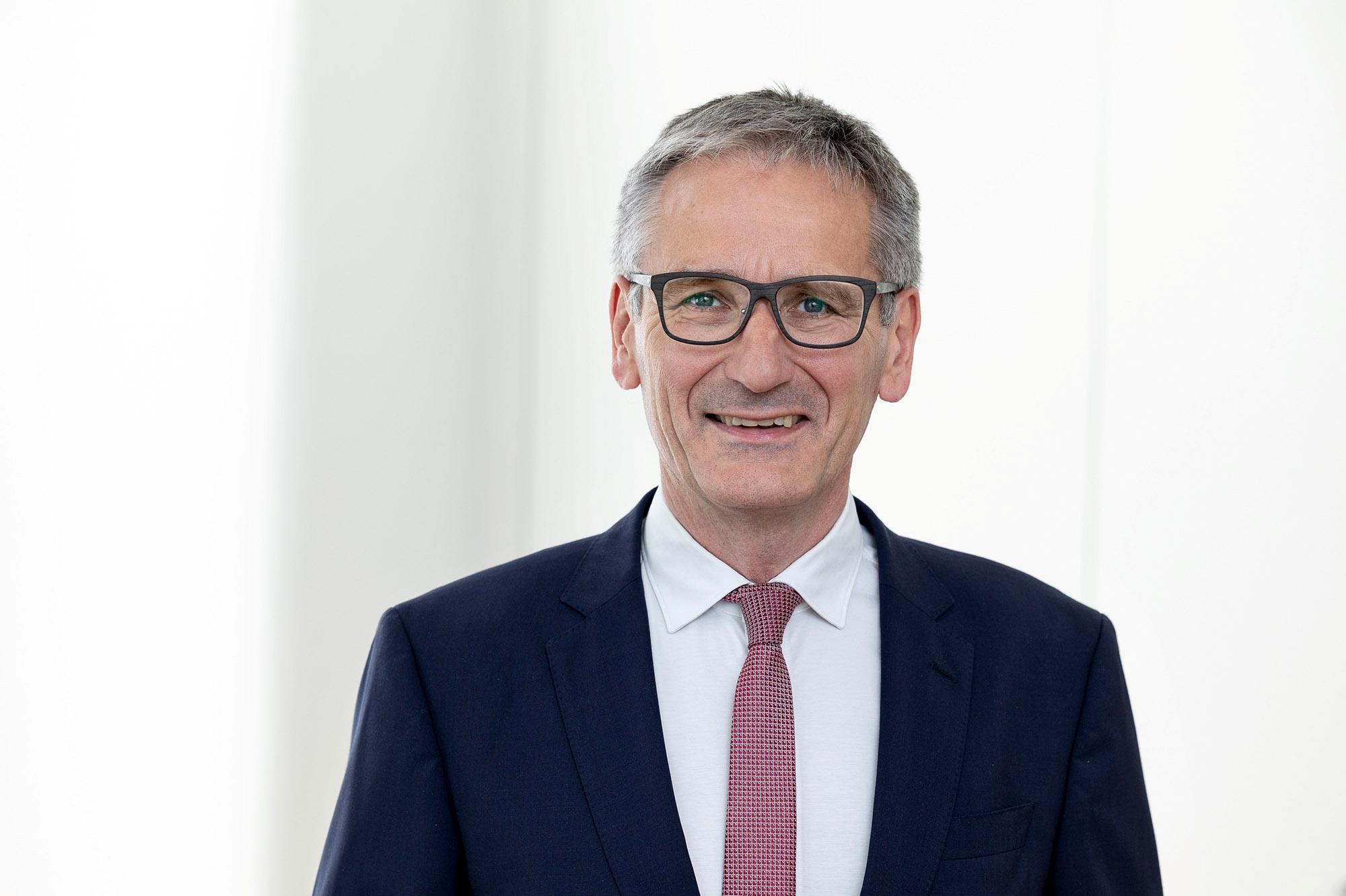 Mann in dunklem Anzug vor hellem Hintergrund blickt geradeaus in die Kamera.