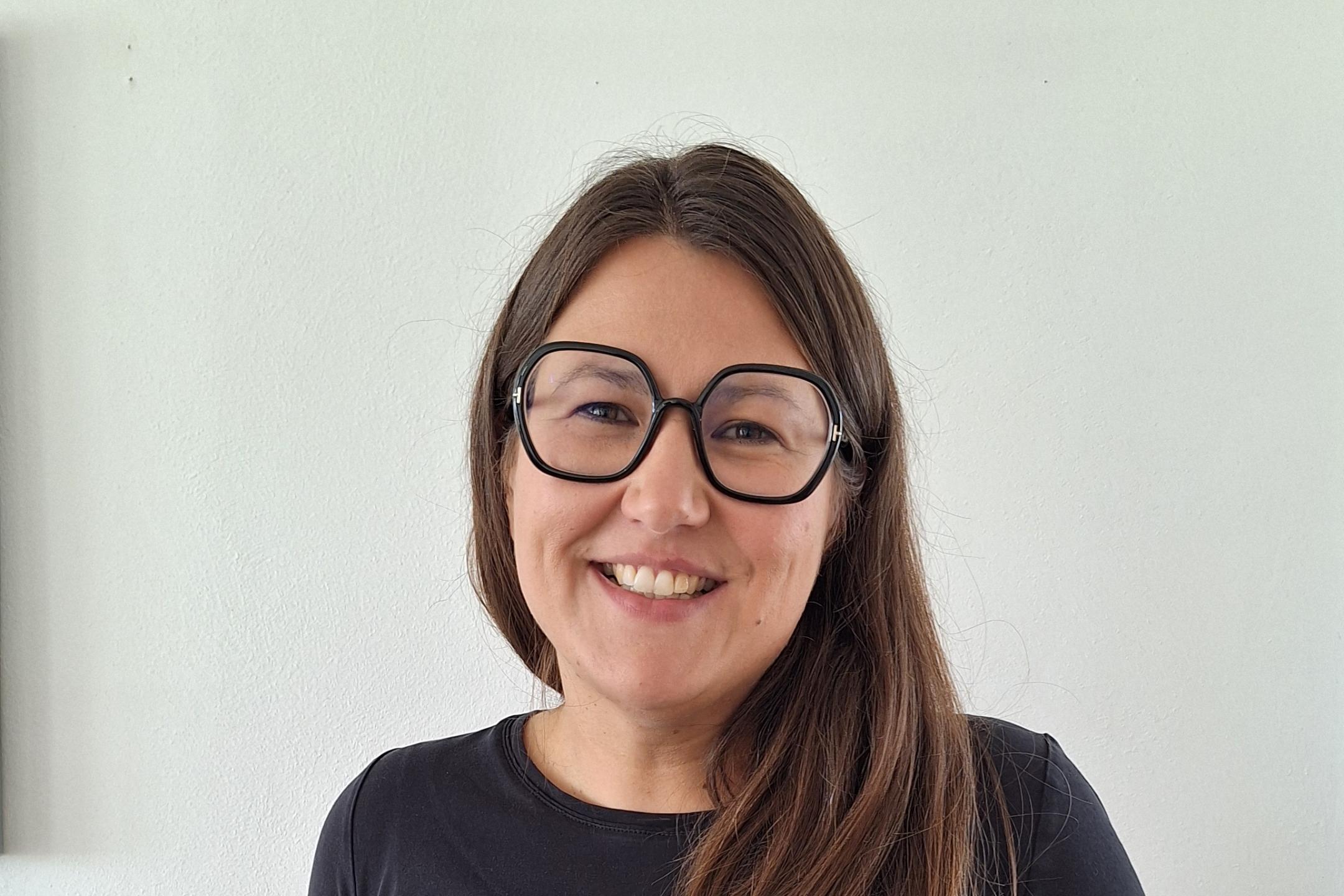Junge Frau mit langen Haaren, runder Brille und T Shirt steht vor weißer Wand.