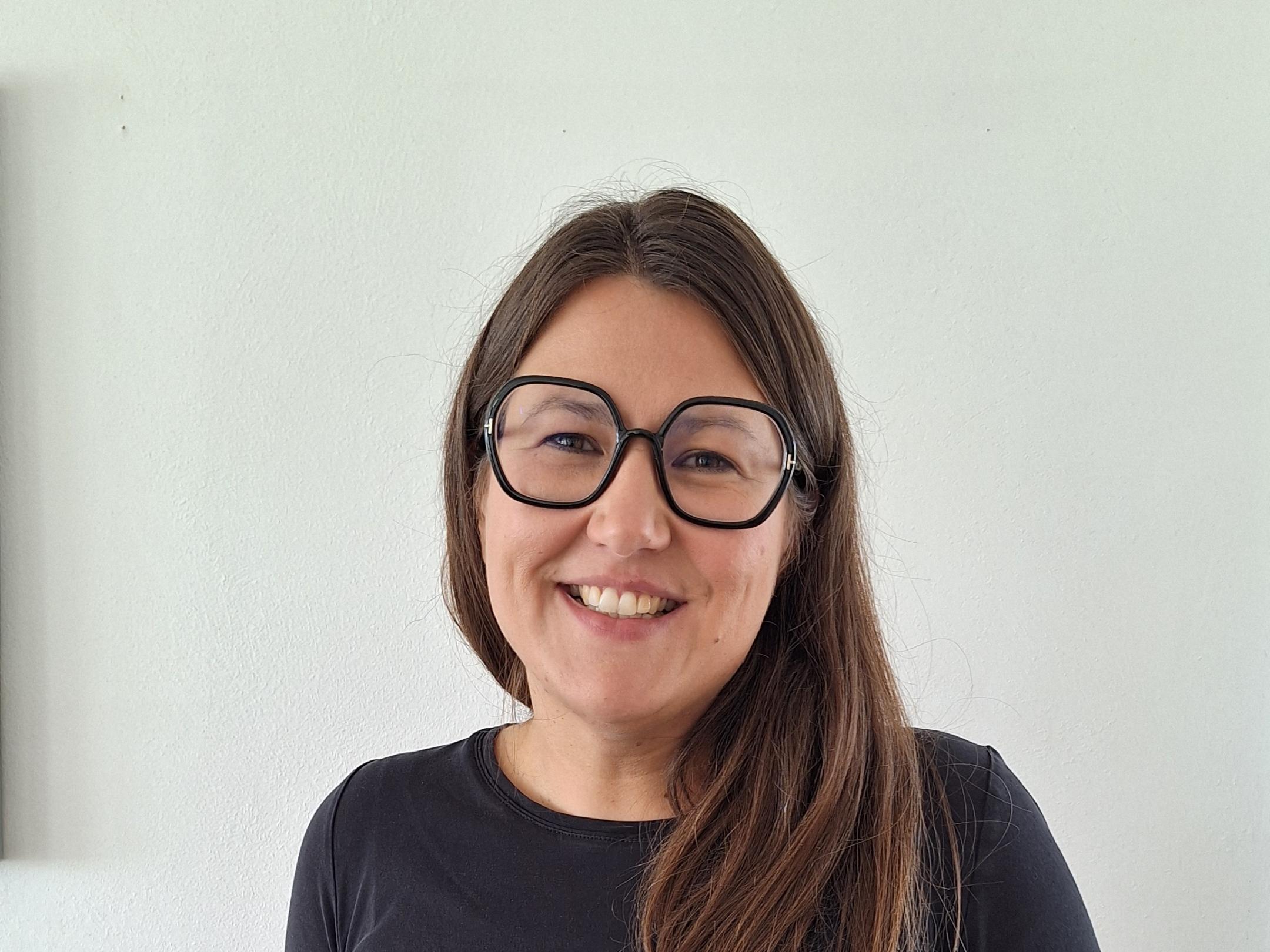 Junge Frau mit langen Haaren, runder Brille und T Shirt steht vor weißer Wand.