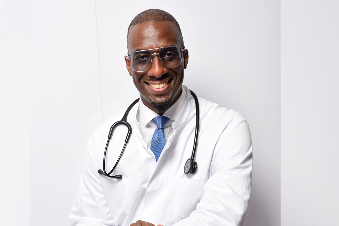Dunkelhäutiger Mann im weißen Arztkittel mit Stethoskop, sitzend vor heller Wand