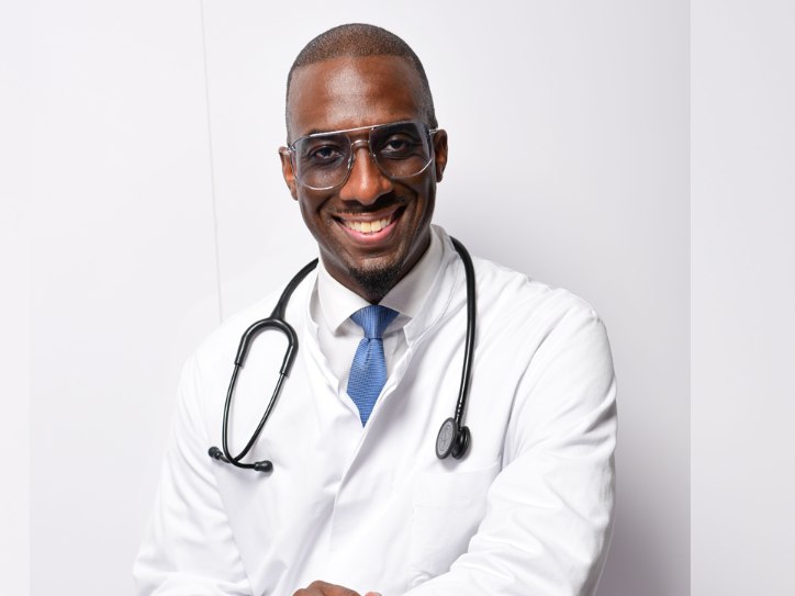 Dunkelhäutiger Mann im weißen Arztkittel mit Stethoskop, sitzend vor heller Wand