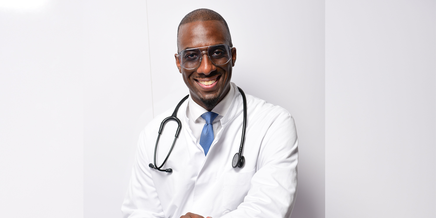 Dunkelhäutiger Mann im weißen Arztkittel mit Stethoskop, sitzend vor heller Wand