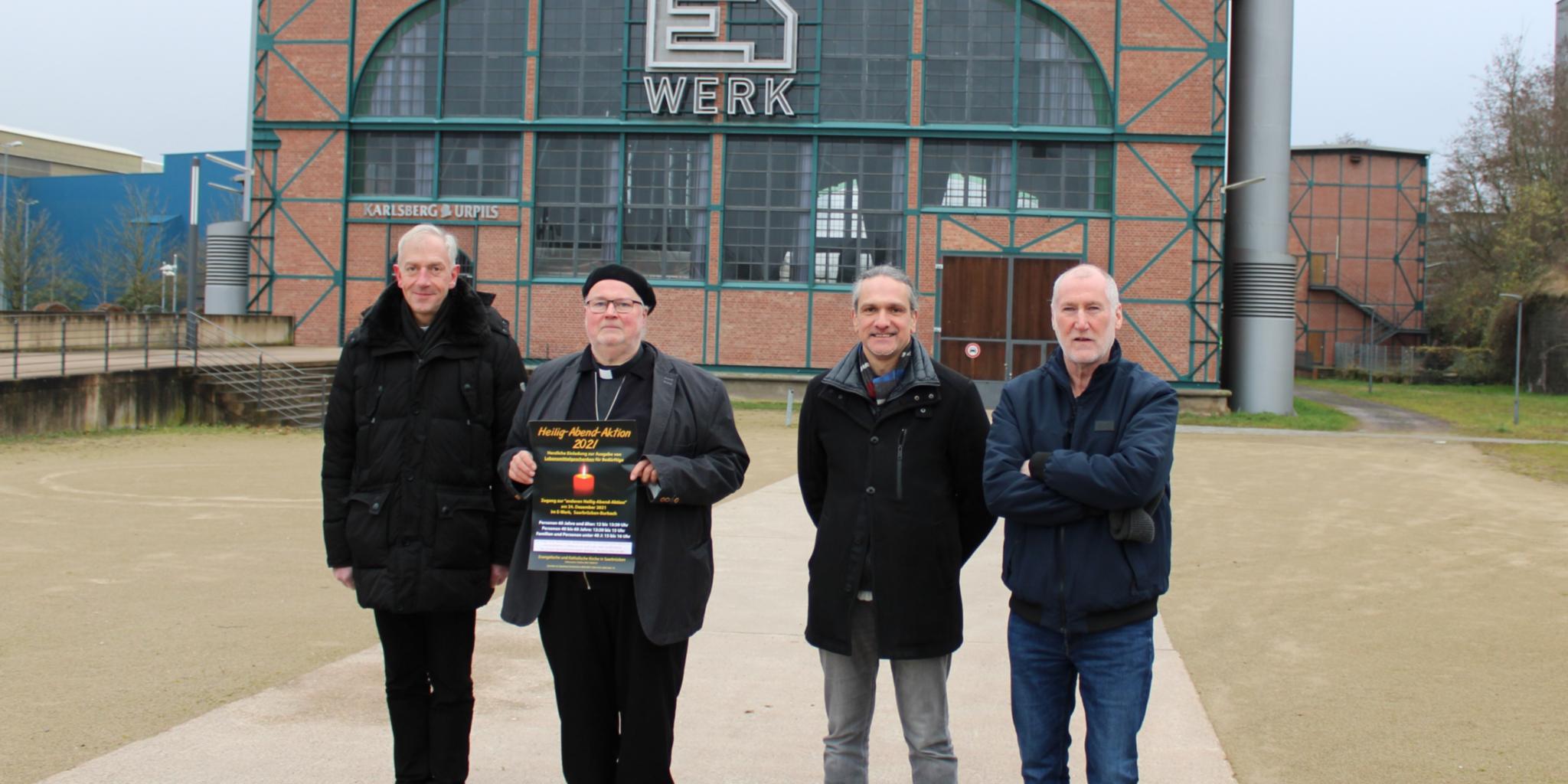 Dechant Benedikt Welter (kth. Dekanat Saarbrücken), Diakon Horst-Peter Rauguth (Organisationsteam Heiligabend-Aktion), Pfarrer Joachim Wörner (Ev. Kirchengemeinde St. Johann / Kirchenkreis Saar-West) und Pastoralreferent Heiner Buchen (kth. Dekanat Saarbrücken) vor dem E-Werk, in dem die Kirchen an Heiligabend wieder eine Aktion für Einsame und Bedürftige durchführen werden. Foto: evks/Eulenstein