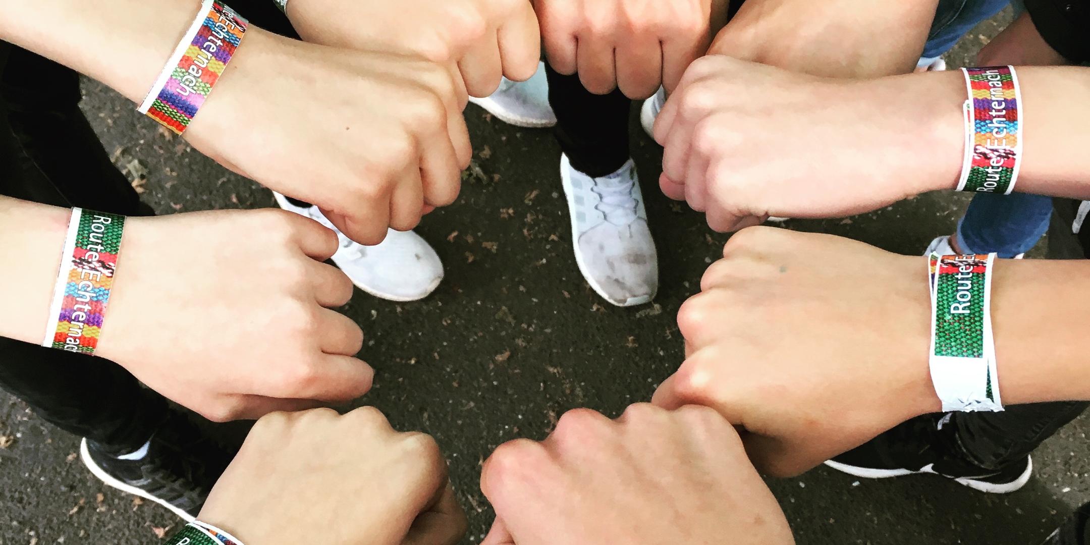 Hände zu lockeren Fäusten geballt, formen sich zu einem Kreis. Alle tragen ein Freundschaftsband, auf dem Route Echternach steht.