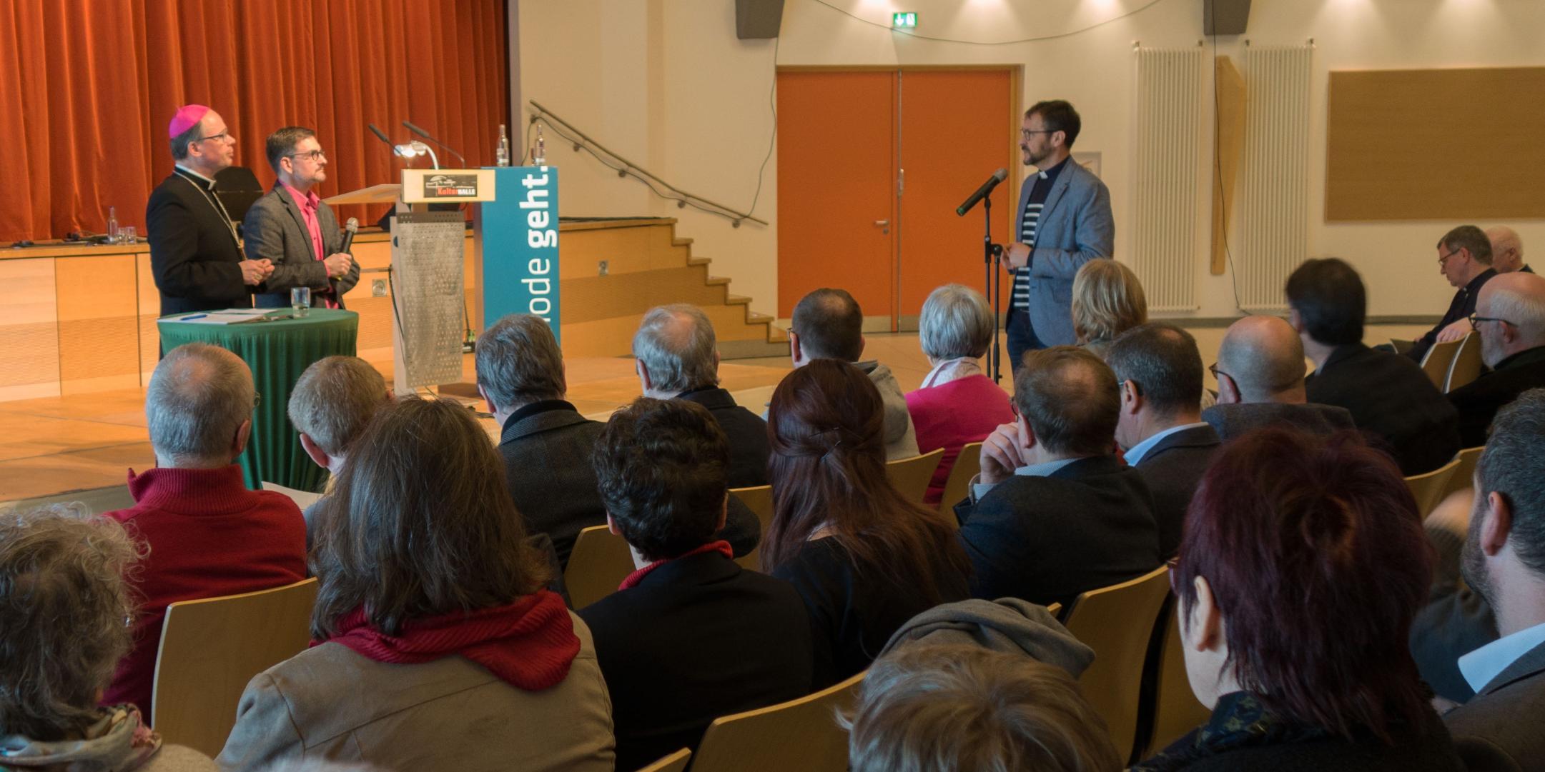 Dechant Jörg Meyrer (rechts) aus Bad Neuenahr-Ahrweiler stellt Bischof Ackermann (links) und Christian Heckmann, Leiter des Synodenbüros, Fragen zur Synodenumsetzung. Viele Leute sitzen im Raum und hören zu.