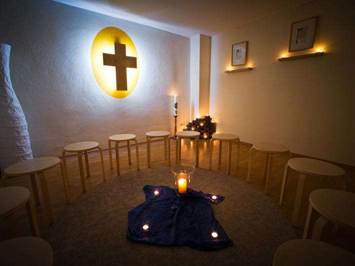 Holzhocker stehen im Kreis, in der Mitte ein blaues Tuch mit Kerze und vier Teelichtern, an der Wand hängt ein Kreuz