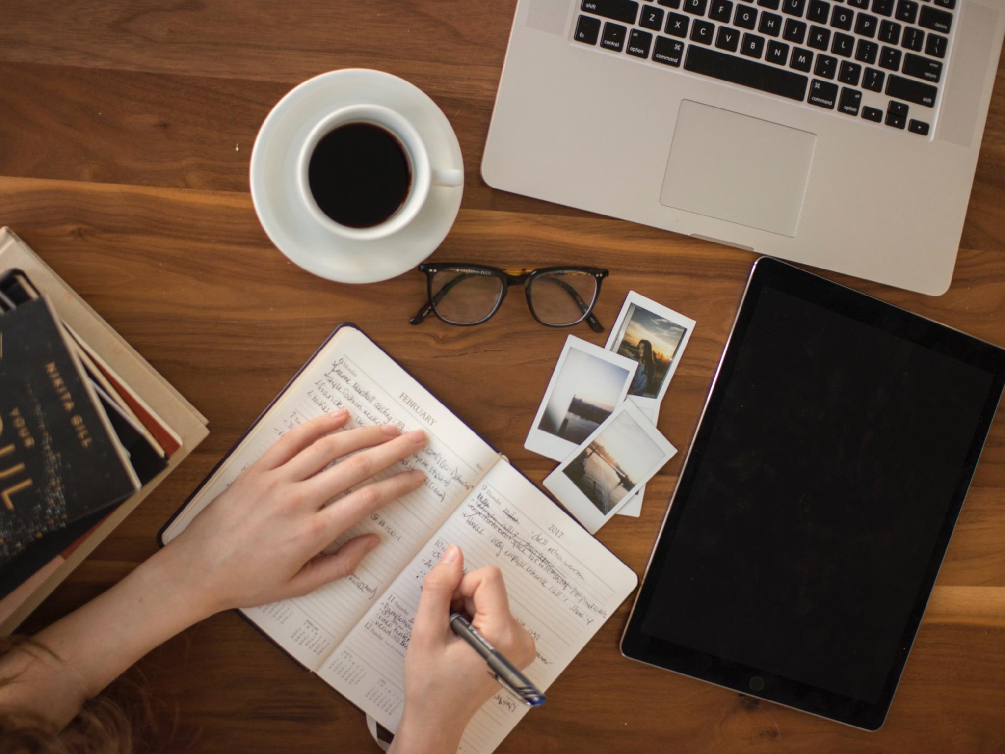 Eine Person schreibt in Notizheft, auf dem Tisch liegen Notebook, Tablet, Brille, Bilder, Kaffeetasse