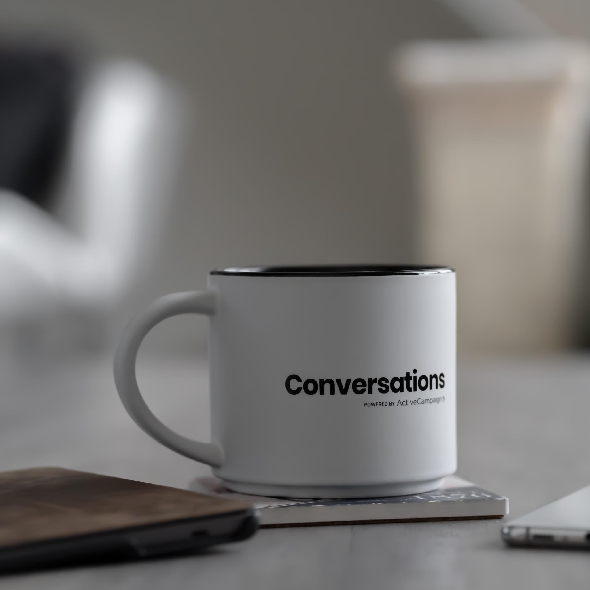 Eine Kaffeetasse mit der Aufschrift converstations steht auf einem Tisch, daneben ein Handy und ein Notebook.